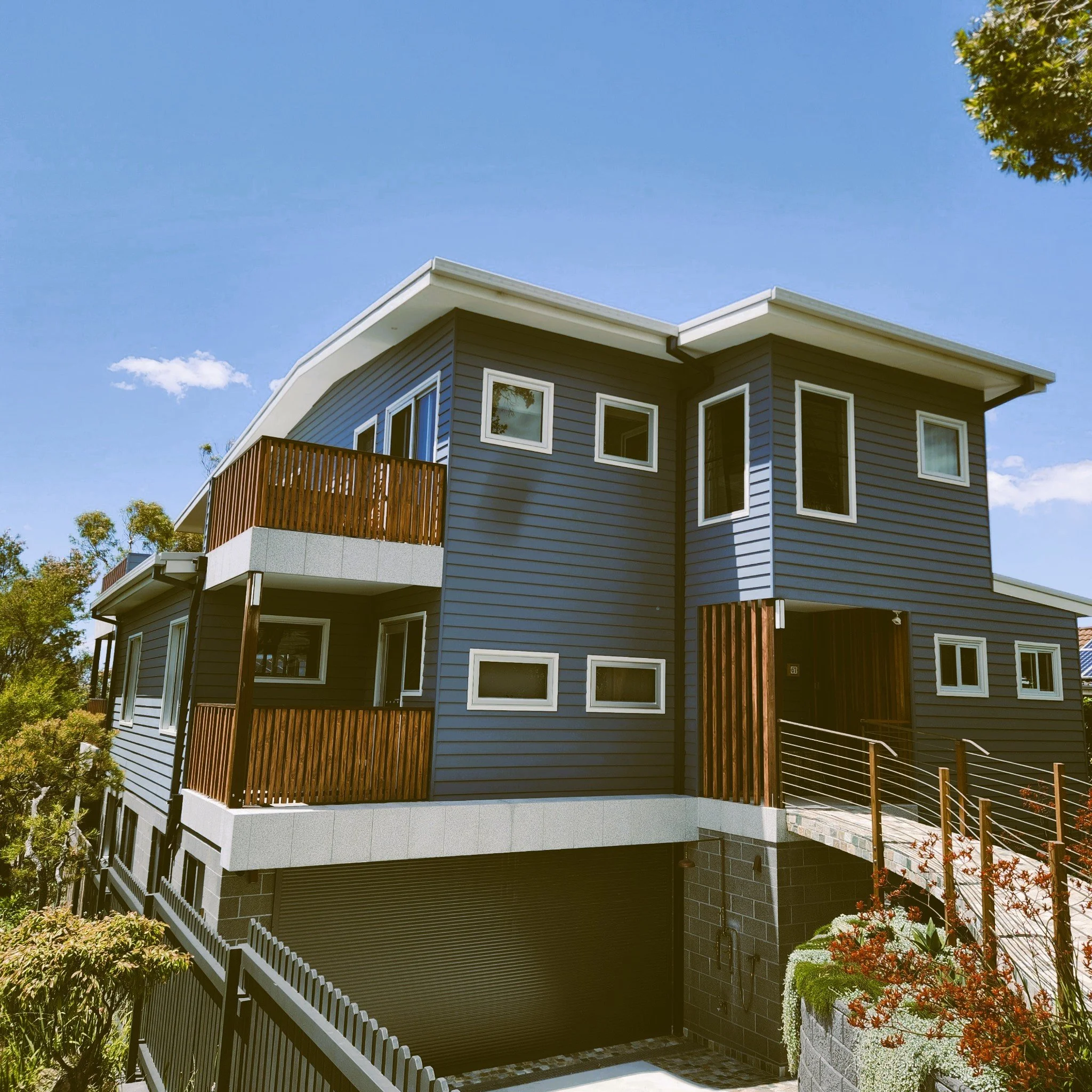 Modern dark blue multi-story house with wooden balconies, multiple windows, and a ramp leading to the entrance, surrounded by greenery under a clear blue sky.