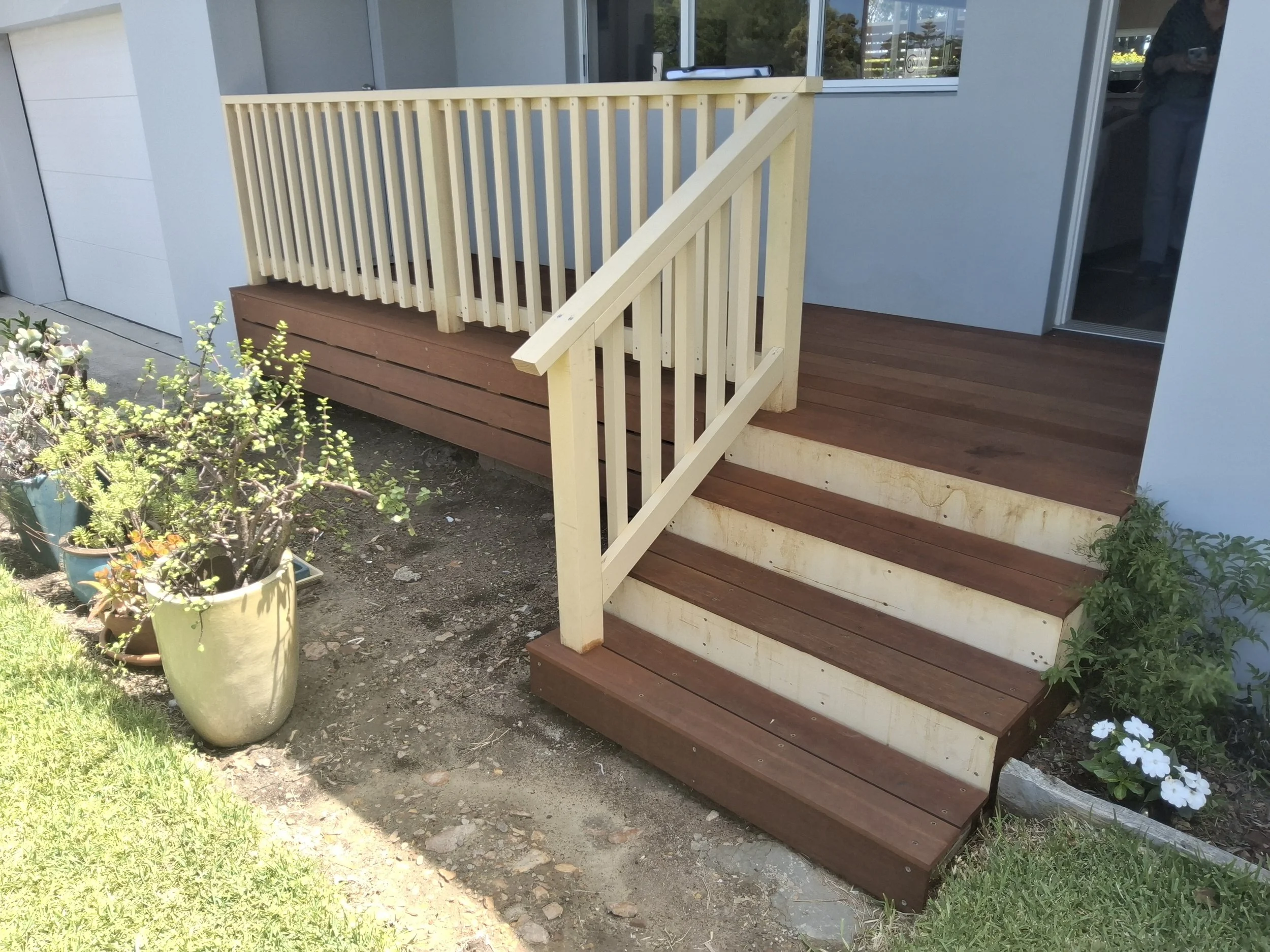 Newly built small wooden deck with stairs and railing attached to a house, with potted plants and garden plants nearby.