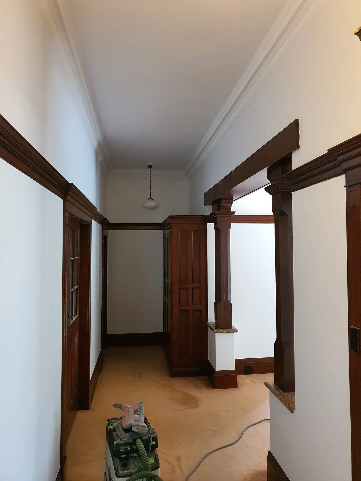 Interior view of a hallway with wooden trim and columns, painting in progress, with a small vacuum or cleaning device on the carpet.