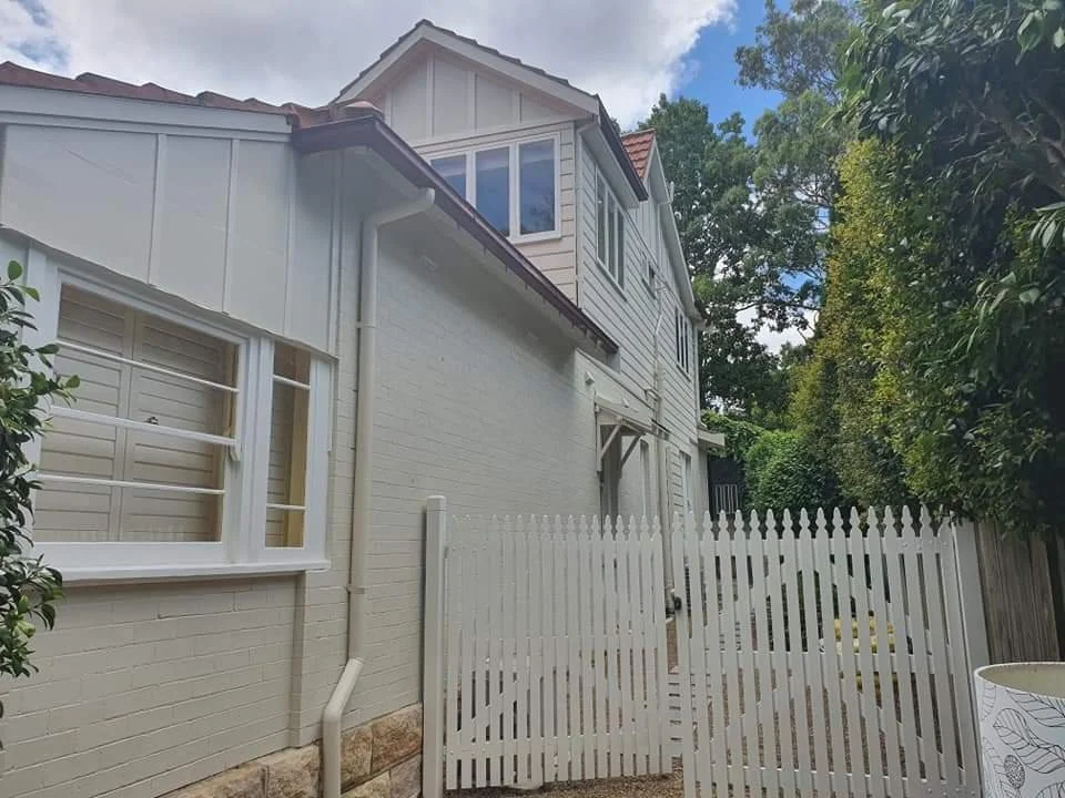 A white brick house with a white picket gate, two-story with multiple windows, surrounded by green trees and bushes.