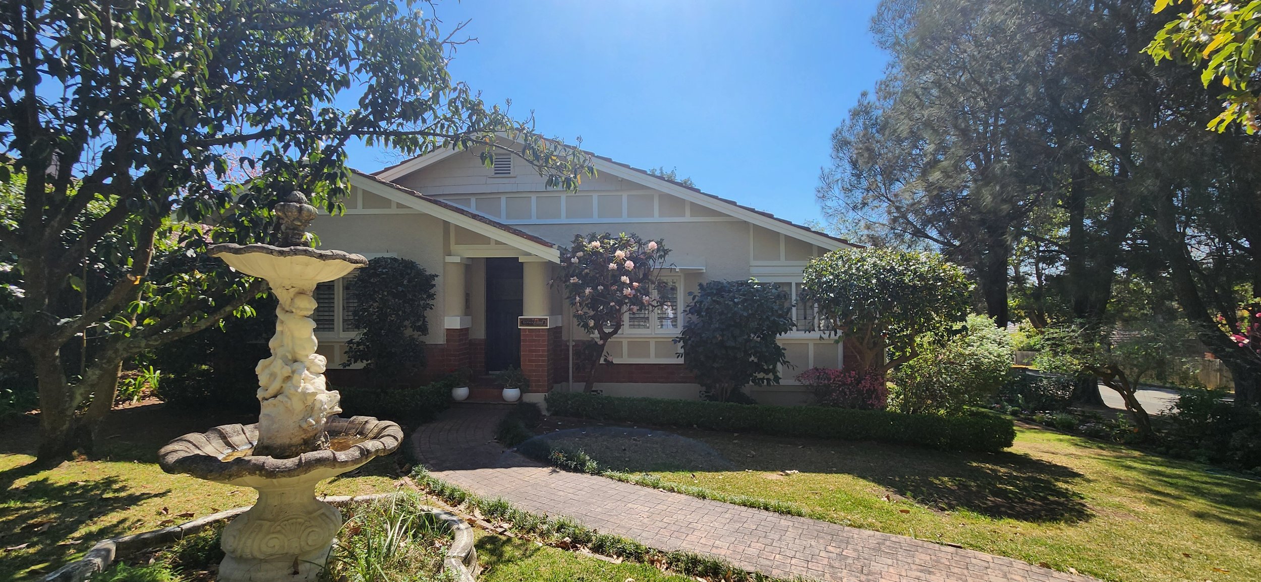 Front yard of a house with a brick and white exterior, surrounded by trees, bushes, and a curved brick pathway. There is a stone fountain in the yard. The sky is clear and sunny.