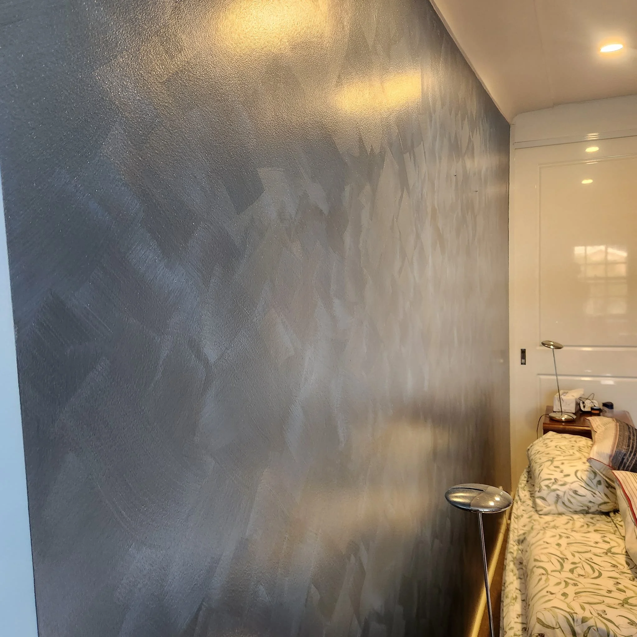 Interior of a bedroom with a textured gray wall, a bed with floral bedding, a nightstand, and a modern reading lamp.