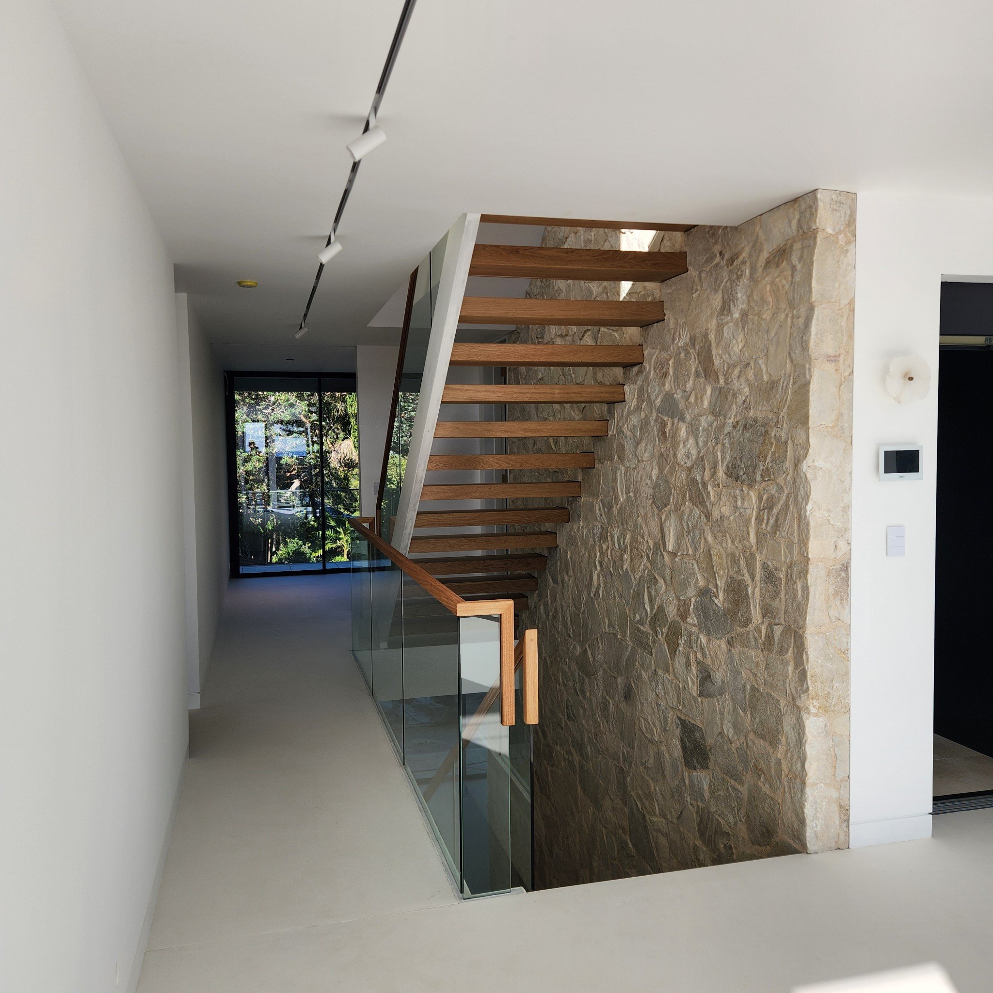 Modern interior staircase with wooden steps, glass railing, and stone wall, in a bright minimalist home with large windows.