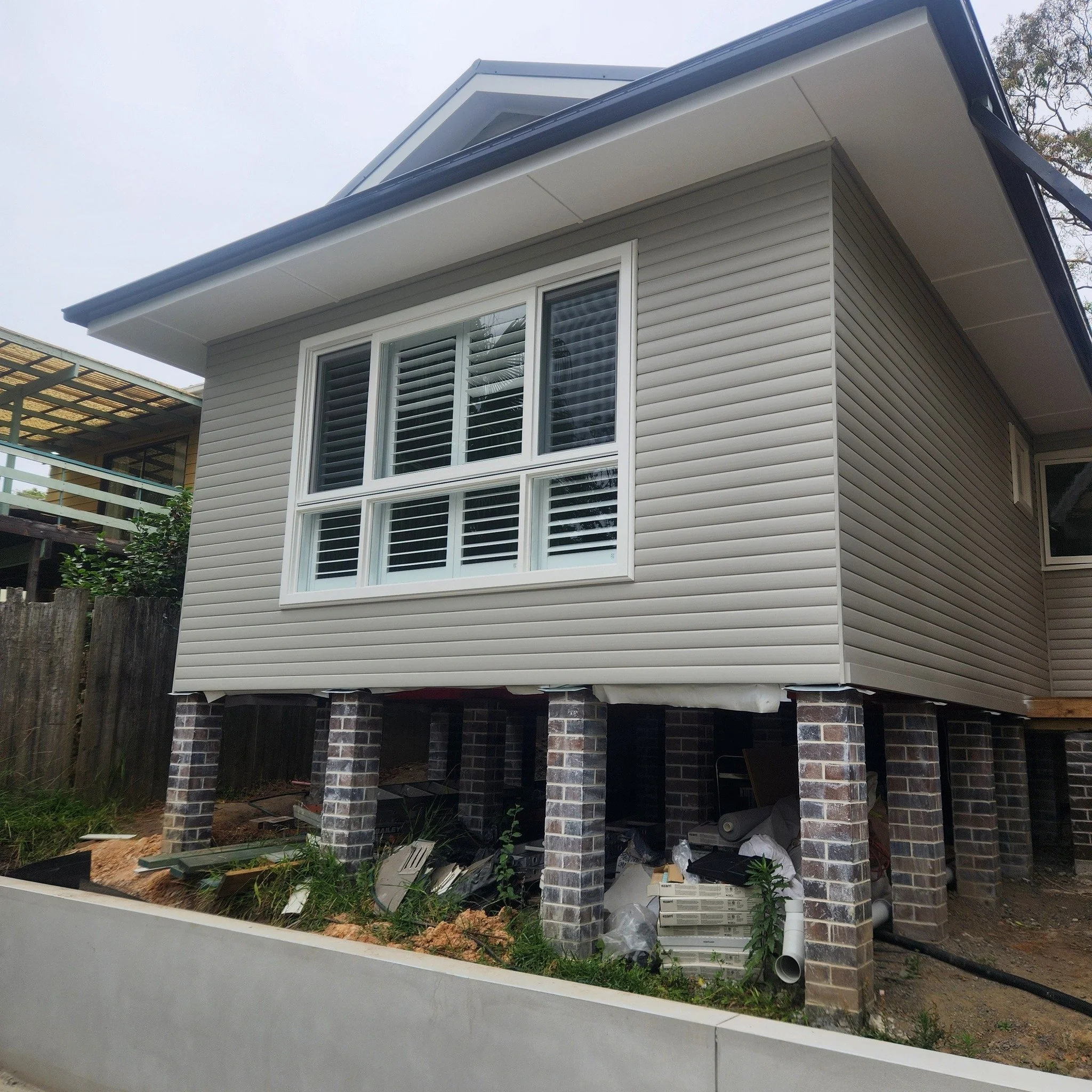 A house under construction with the exterior siding partially installed, elevated on brick pillars, with construction debris and materials underneath.