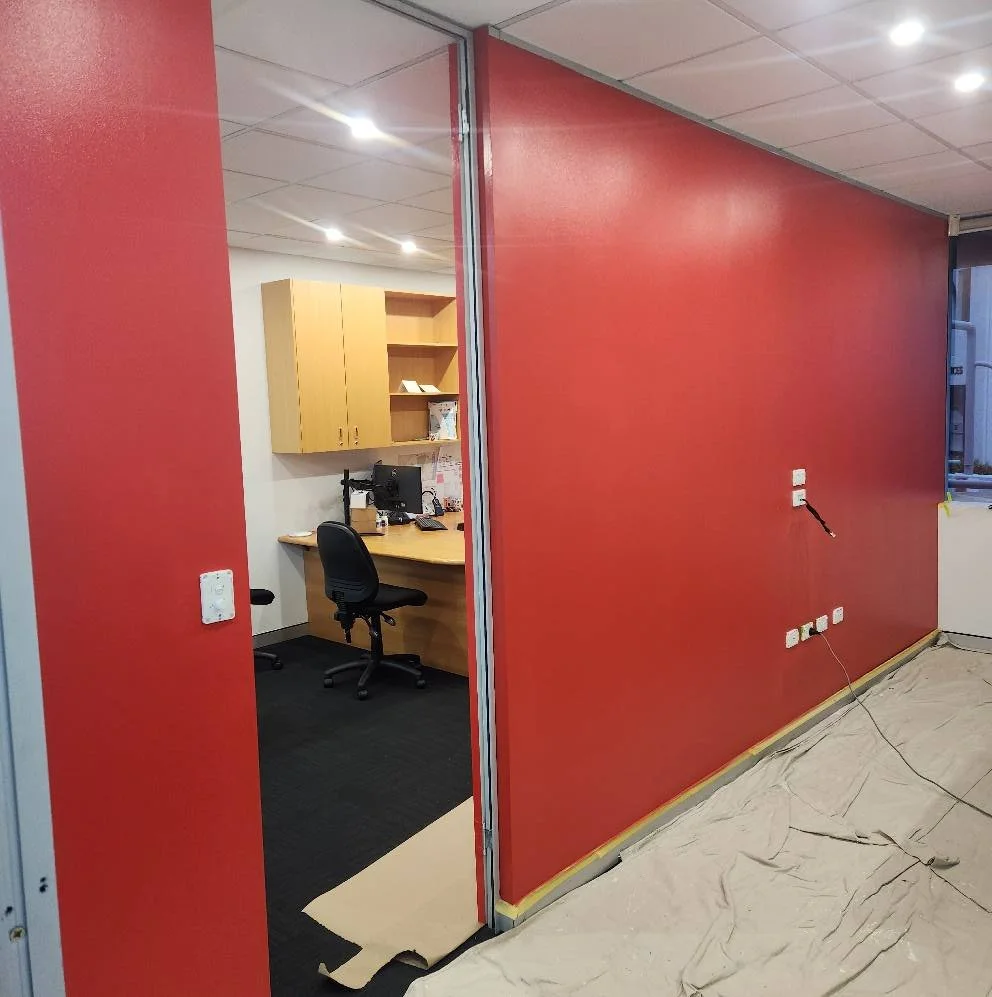 An office space with a red wall and a doorway leading to a small office area with a wooden desk, a black office chair, and yellow cabinets. The floor in the main area is covered with paper, and multiple electrical outlets and wires are visible on the red wall.
