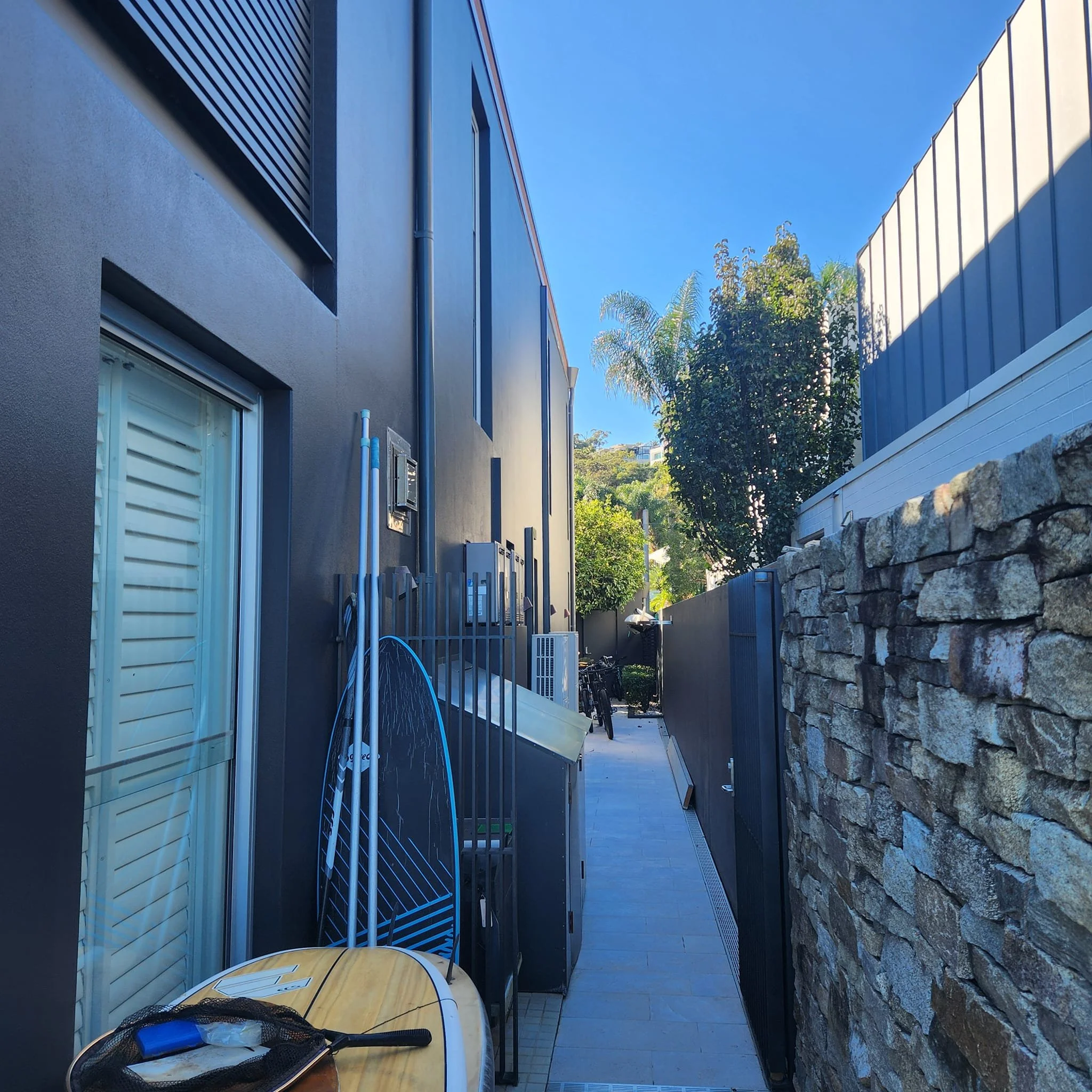 Narrow outdoor alley between a modern black building and a stone wall, with bicycles, outdoor equipment, and trees in the background.