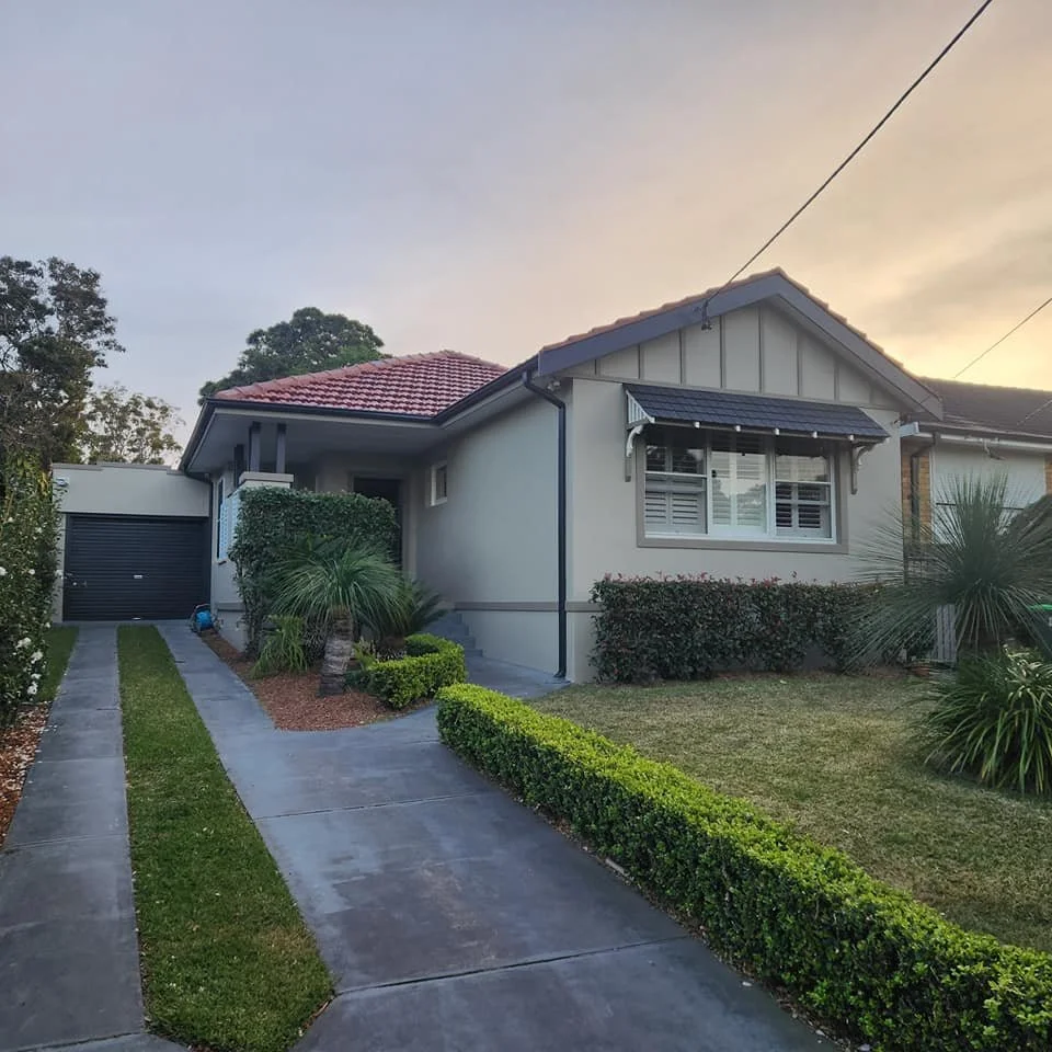 A modern suburban house with white exterior walls, a red-tiled roof, and a small front yard with trimmed bushes and palm trees. A concrete driveway leads to a black garage door.