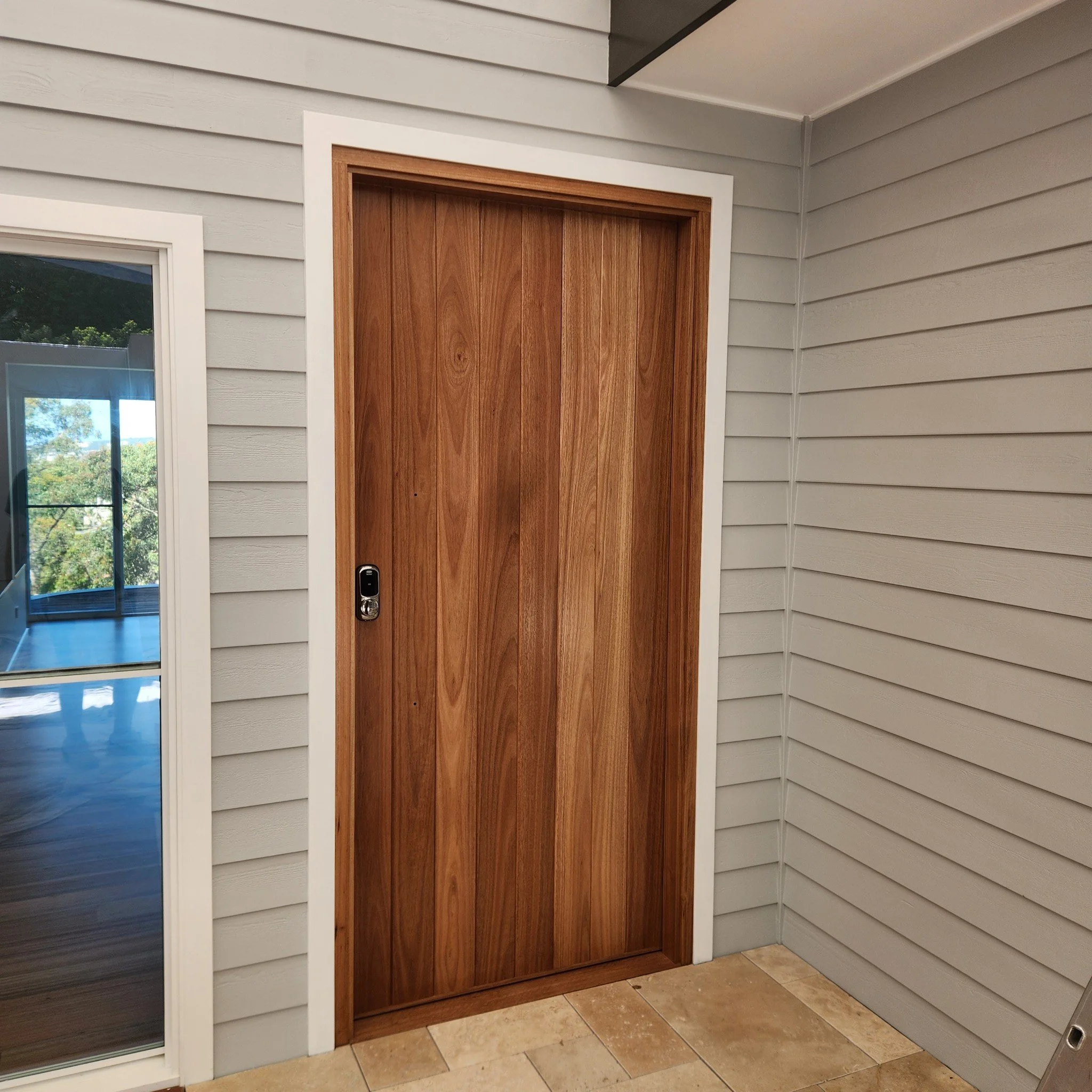 A wooden sliding door in an interior space, with a white frame around it, situated between gray siding walls and next to a glass door leading to an outdoor area.