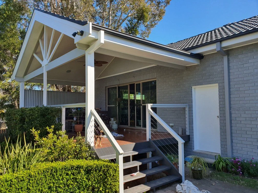 View of a house patio with steps leading up to a sliding glass door, gray brick wall, and a white door. The patio has a roof with wooden beams, a ceiling fan, and some potted plants, surrounded by greenery and shrubs.