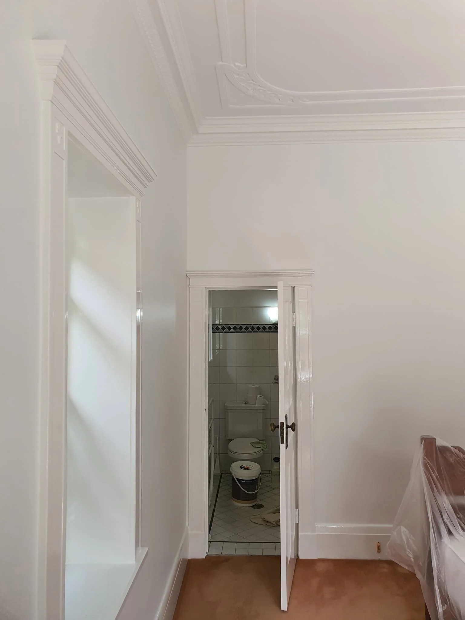 A room with white walls and decorative crown molding, open door leading to a small bathroom with a toilet, partially visible shower, and some cleaning supplies on the floor.
