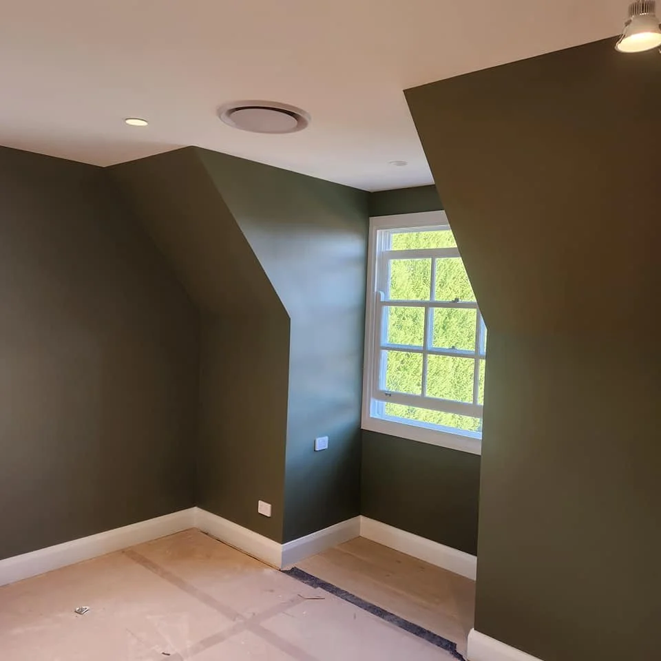 Empty room with green walls, a window showing greenery outside, and newly painted white baseboards.
