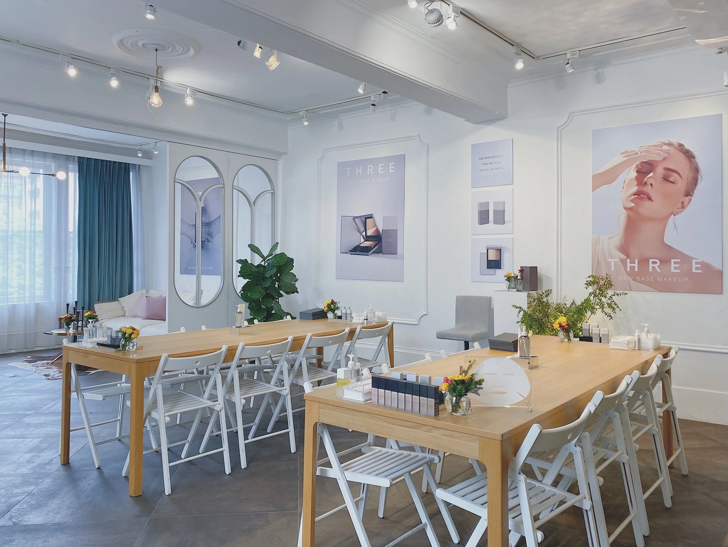 A modern beauty or makeup studio with light wooden tables, white chairs, large posters advertising makeup products, and decorative plants. There is a seating area with teal curtains and a window.