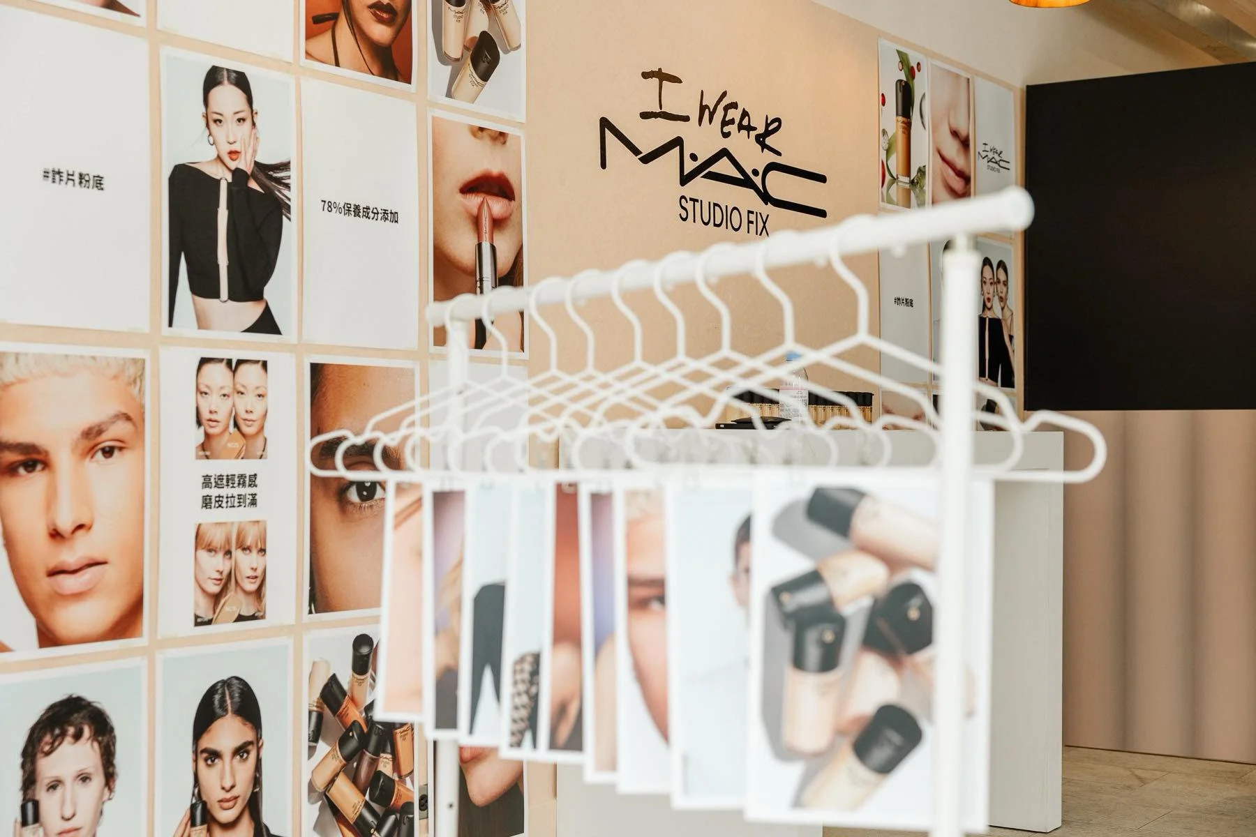 Wall with posters advertising makeup products, including models and close-up images of lips and eyes, with a MAC Cosmetics logo and the text "I Wear MAC Studio Fix." In the foreground, a white rack displays samples of makeup bottles and brushes.