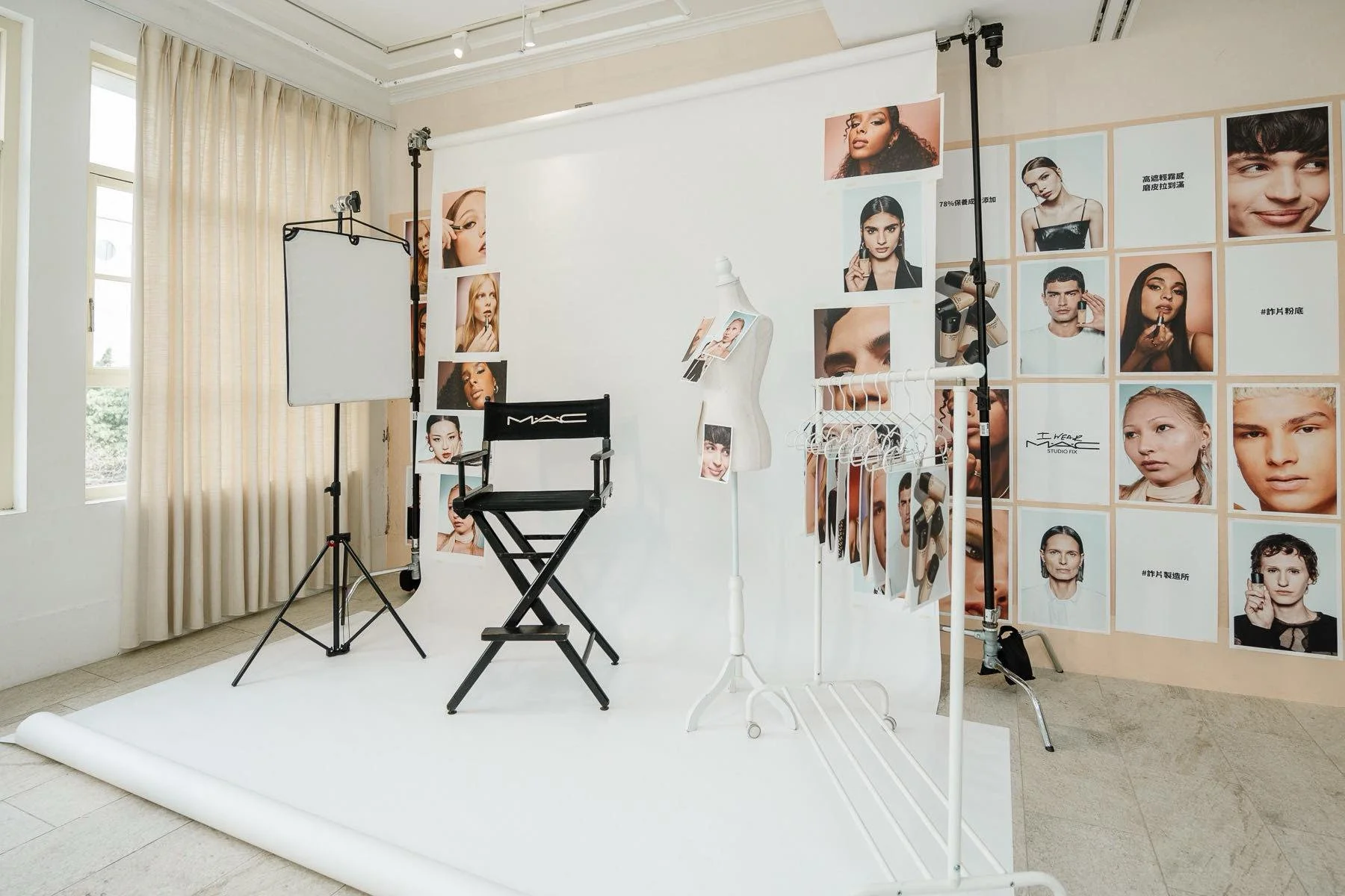 Photography studio setup with a white backdrop, studio lights, a director's chair labeled 'MAC', a mannequin with makeup photos, a rack with makeup brushes and tools, and mood board or photo collage of diverse models on the wall.