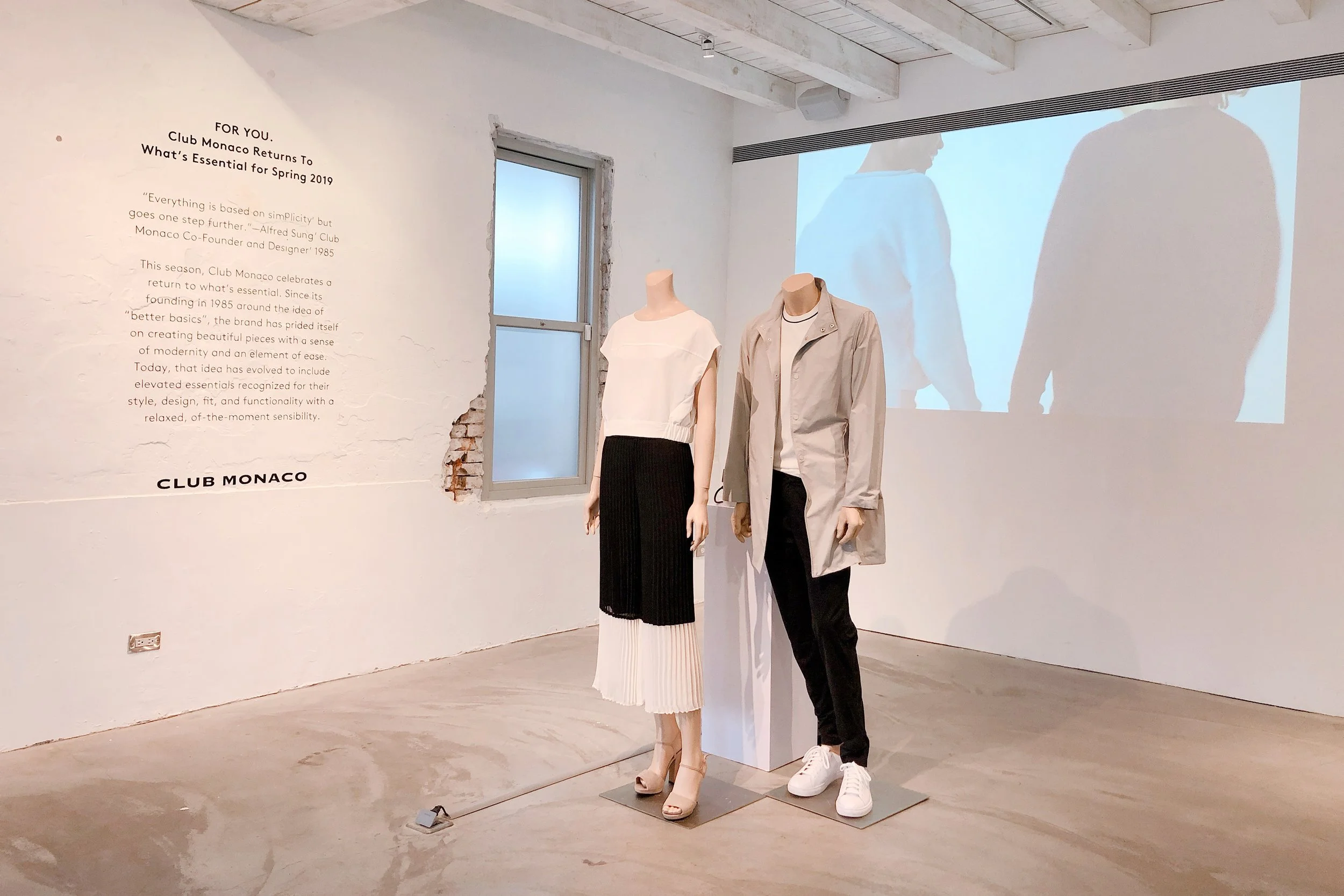 Two mannequins display beige and black clothing in an art gallery with textured white walls, a small window, and a large projection of the mannequins on the wall.