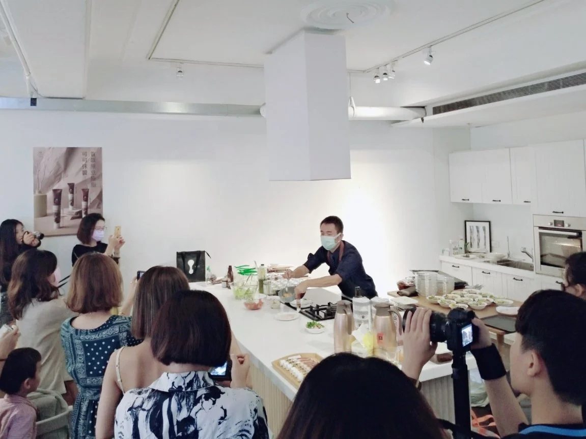 A cooking demonstration with a chef wearing a face mask, surrounded by an audience in a modern kitchen.