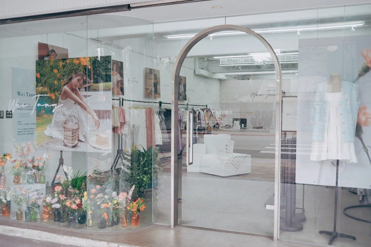 View of a clothing store display window with a floral arrangement, a poster of a girl in a white dress sitting outdoors, and various clothing items inside the store. The store has a modern, minimalist design with white walls and furniture.
