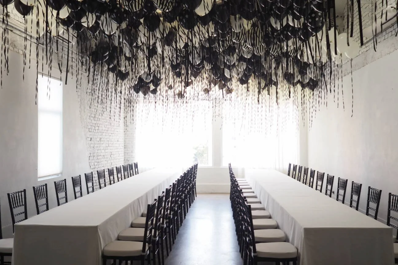 A large, bright conference room with white walls, black chairs, and long white tables arranged in a U-shape. The ceiling is decorated with an artistic installation of hanging black and white paper umbrellas.