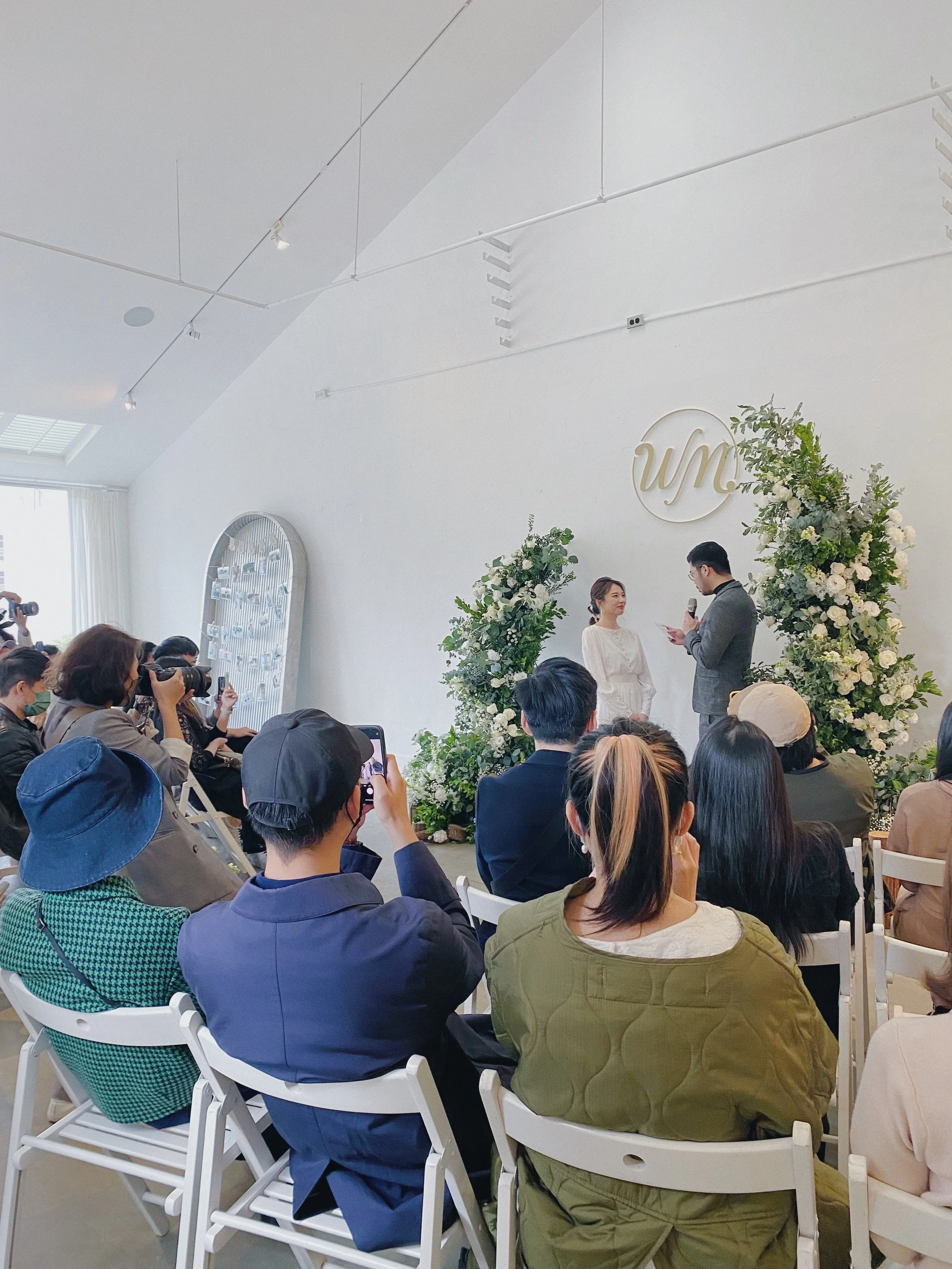 A wedding ceremony with the bride and groom standing at the altar surrounded by floral arrangements, while guests sit and watch, some taking photos. The setting is minimalistic with white walls and a logo on the wall behind them.