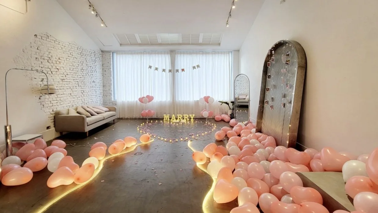 Decorated room for a celebration with pink and white balloons, a "HAPPY" sign with lights, and pink heart-shaped balloons. There are white curtains, a sofa, a mirror, and a banner hanging from the ceiling.
