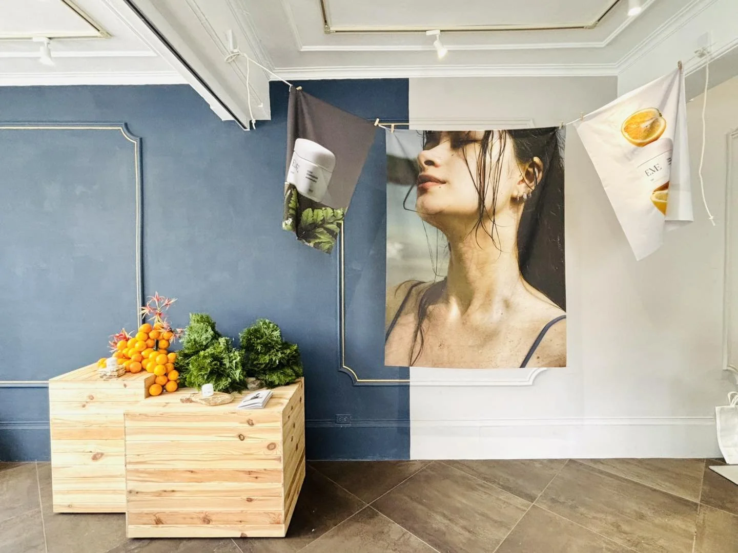 Art installation with hanging fabric banners featuring images of skincare or beauty products, large photograph of a woman with wet hair, wooden display with oranges and greenery, in an indoor gallery or store setting.
