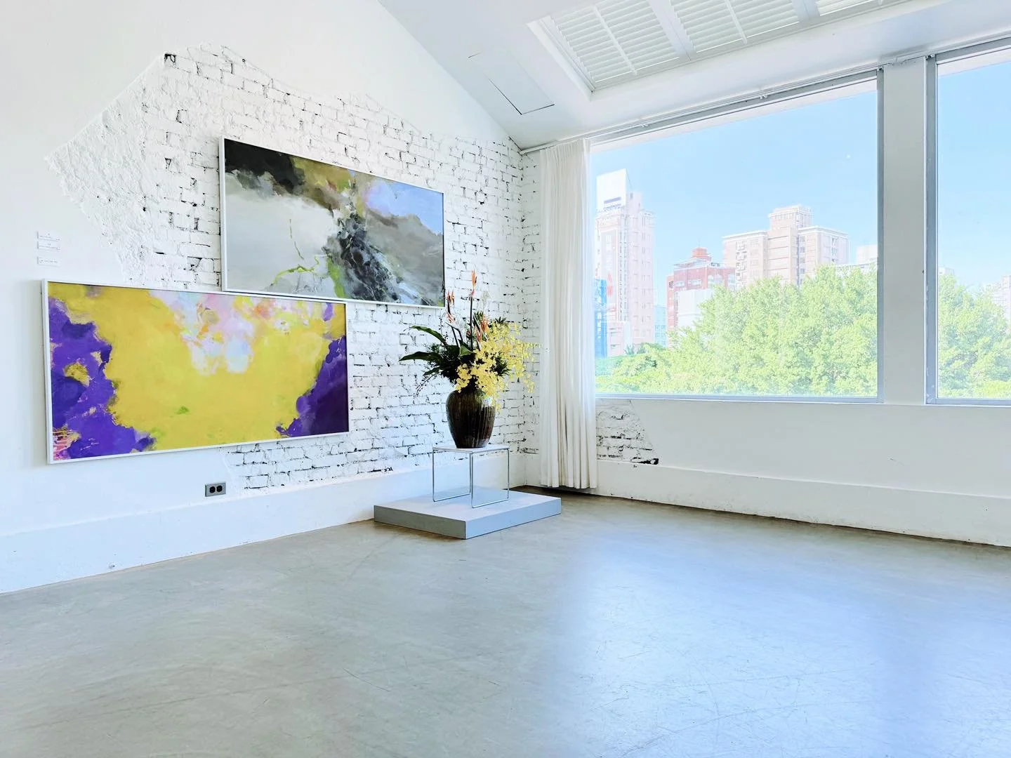 Modern art gallery with two colorful abstract paintings on a white brick wall, a large window with a city view, a tall ceramic vase with flowers on a stand, and natural light filling the room.