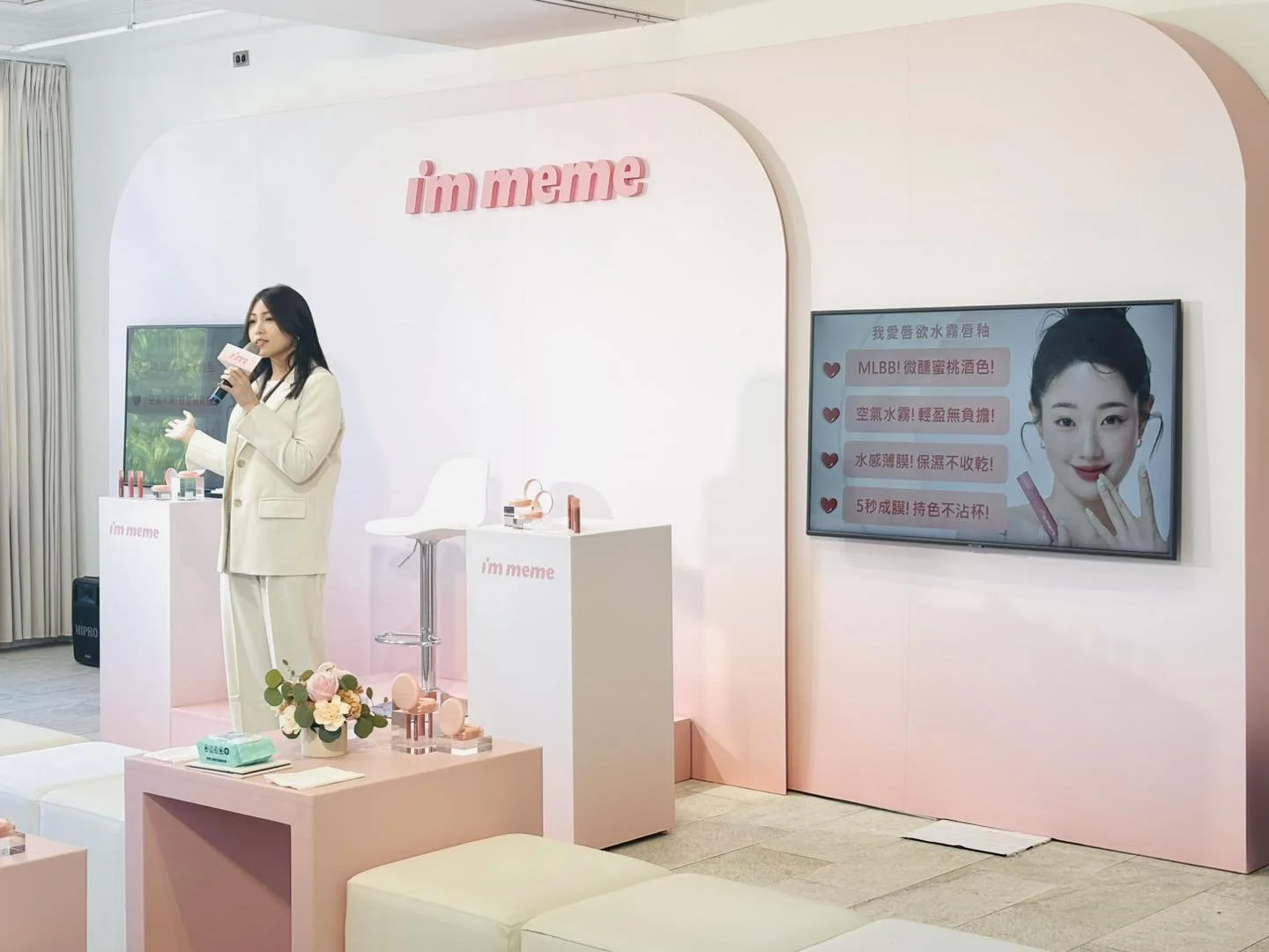 A woman in a white suit presenting at a pink-themed beauty product event called 'I'm meme.' She is holding a microphone, standing behind a pink podium with beauty products. There is a large screen displaying a woman's face with text in Chinese, and t