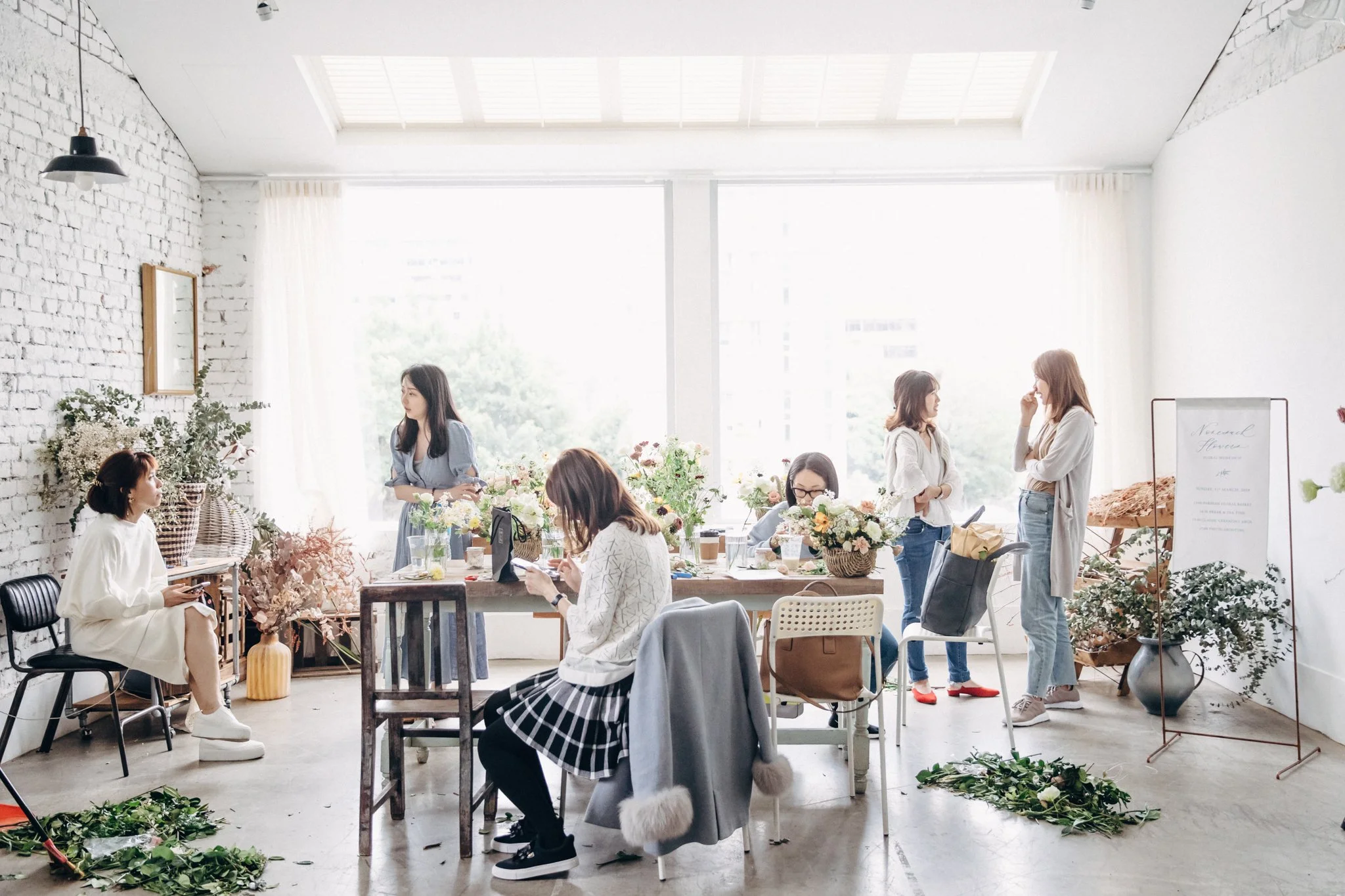 Women preparing floral arrangements in a bright, modern flower shop with white brick walls and large windows.
