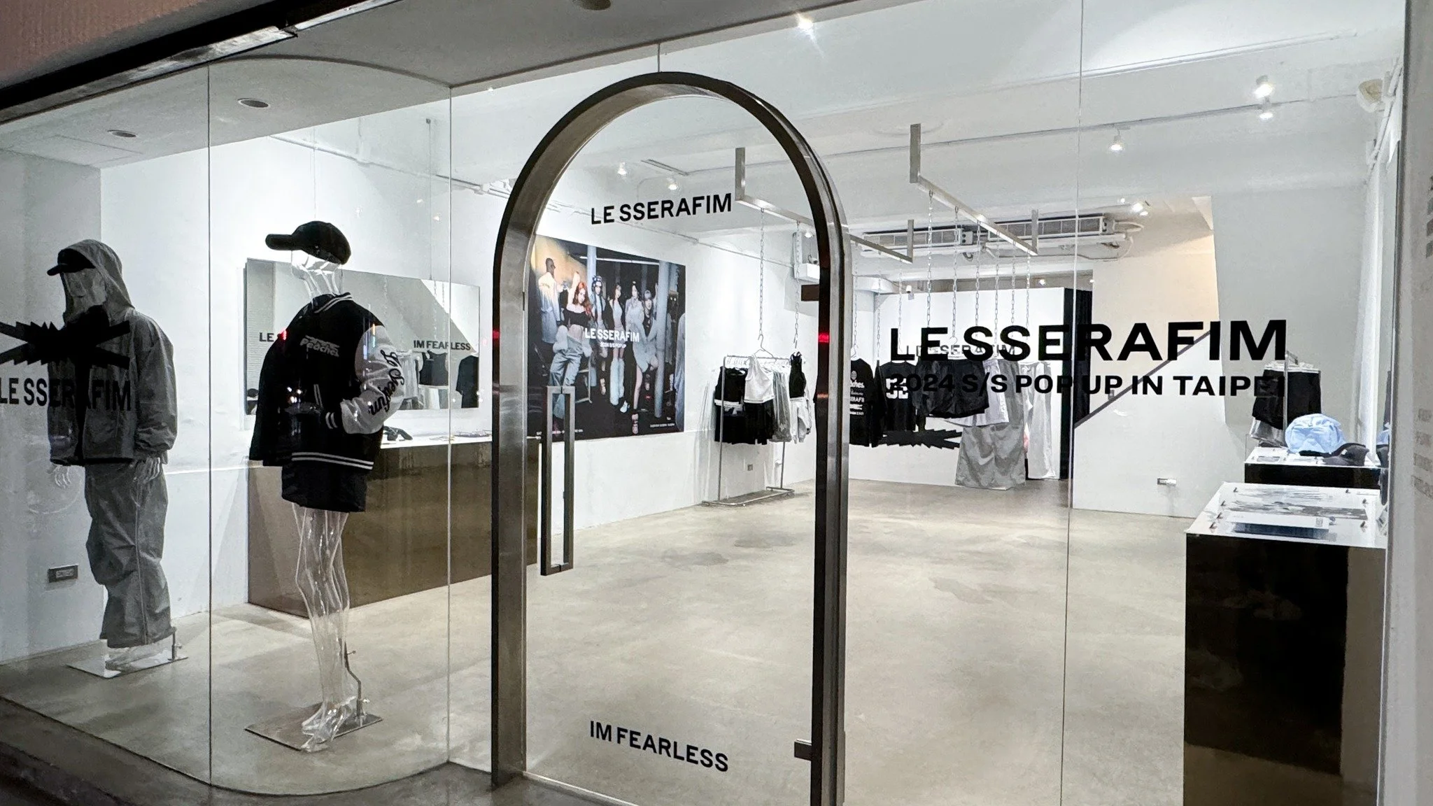 Clothing store interior with mannequins dressed in casual outfits, hanging racks, and promotional poster. Glass storefront with black and white text. Modern minimalistic design with white walls and concrete floor.