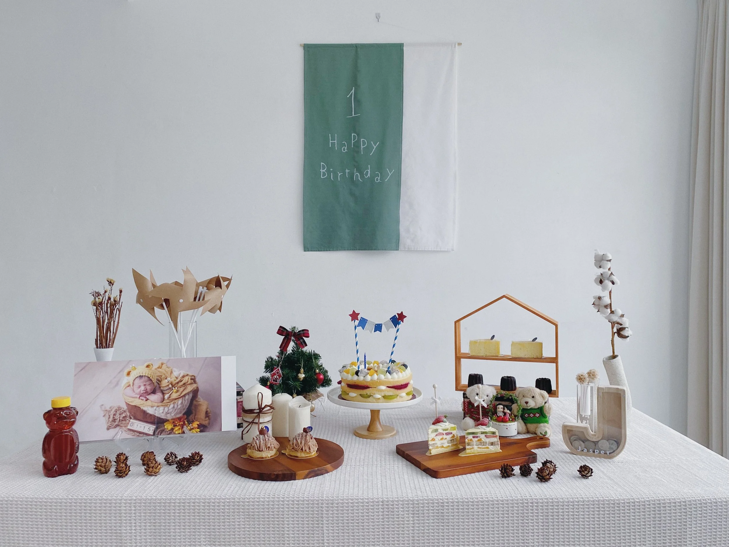 Birthday celebration with a decorated table featuring a cake, balloons, stuffed animals, candles, and pinecones, with a photo of a baby and a small tree and handcrafted items in the background.