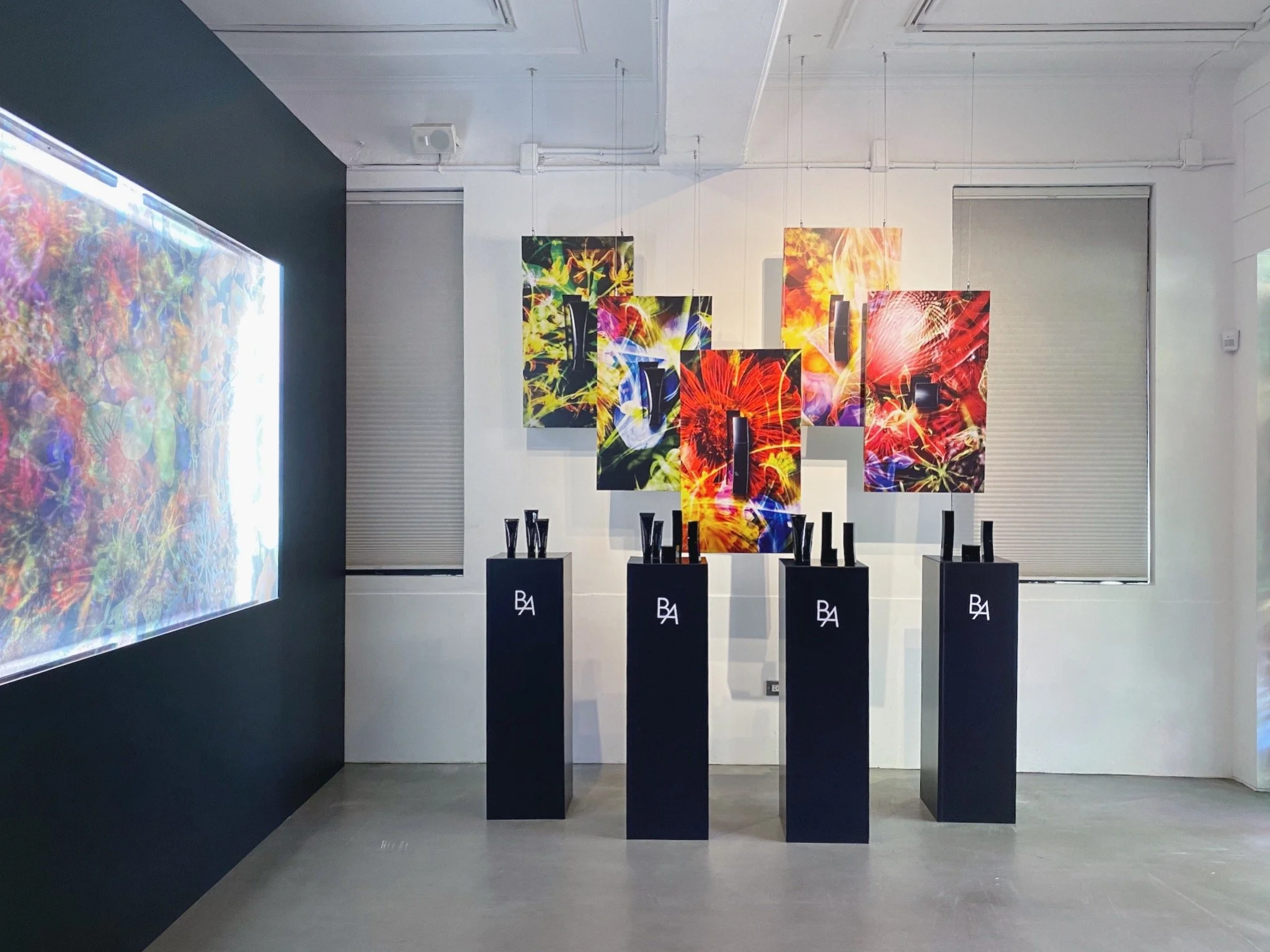 Art gallery display featuring colorful floral artworks mounted on the wall with black pedestals holding black vases or sculptures in front of the artwork.