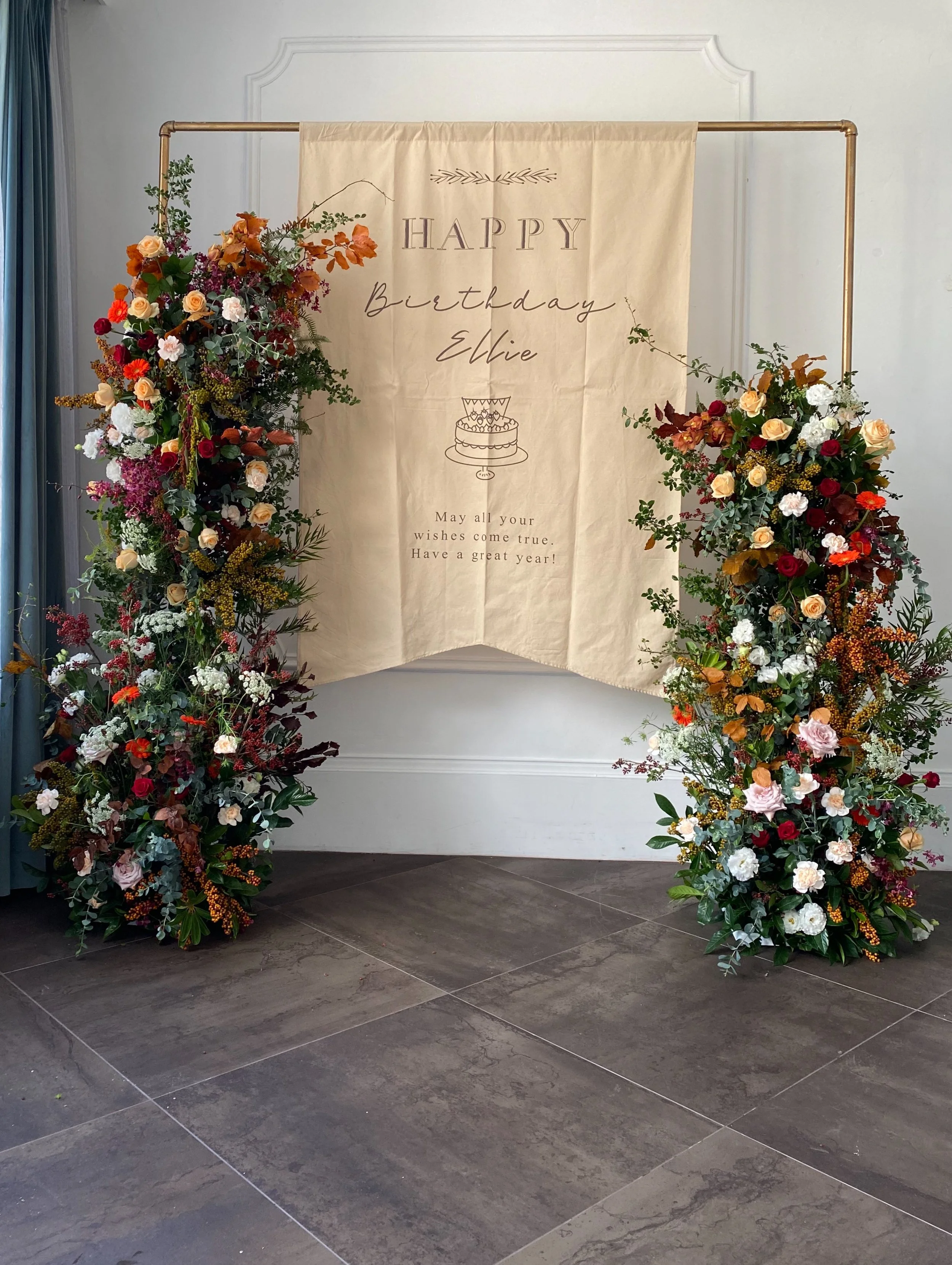Floral arch with a beige banner that reads 'HAPPY Birthday Ellie,' with a cake illustration and a birthday wish. The arch is decorated with assorted colorful flowers and greenery.