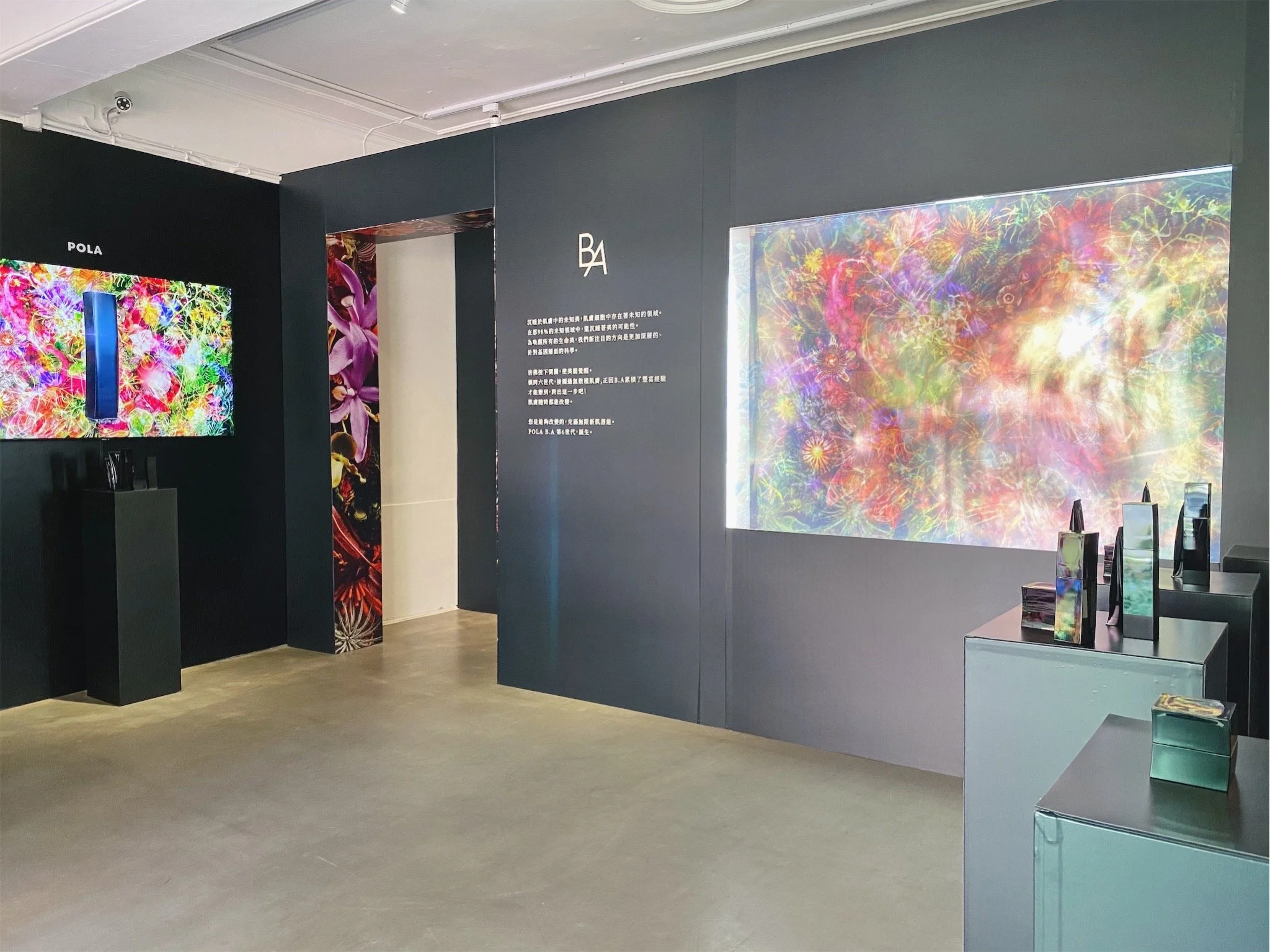 Art gallery exhibition with colorful digital art displays, including a large screen showing abstract floral and cosmic imagery, and smaller holographic devices on pedestals.