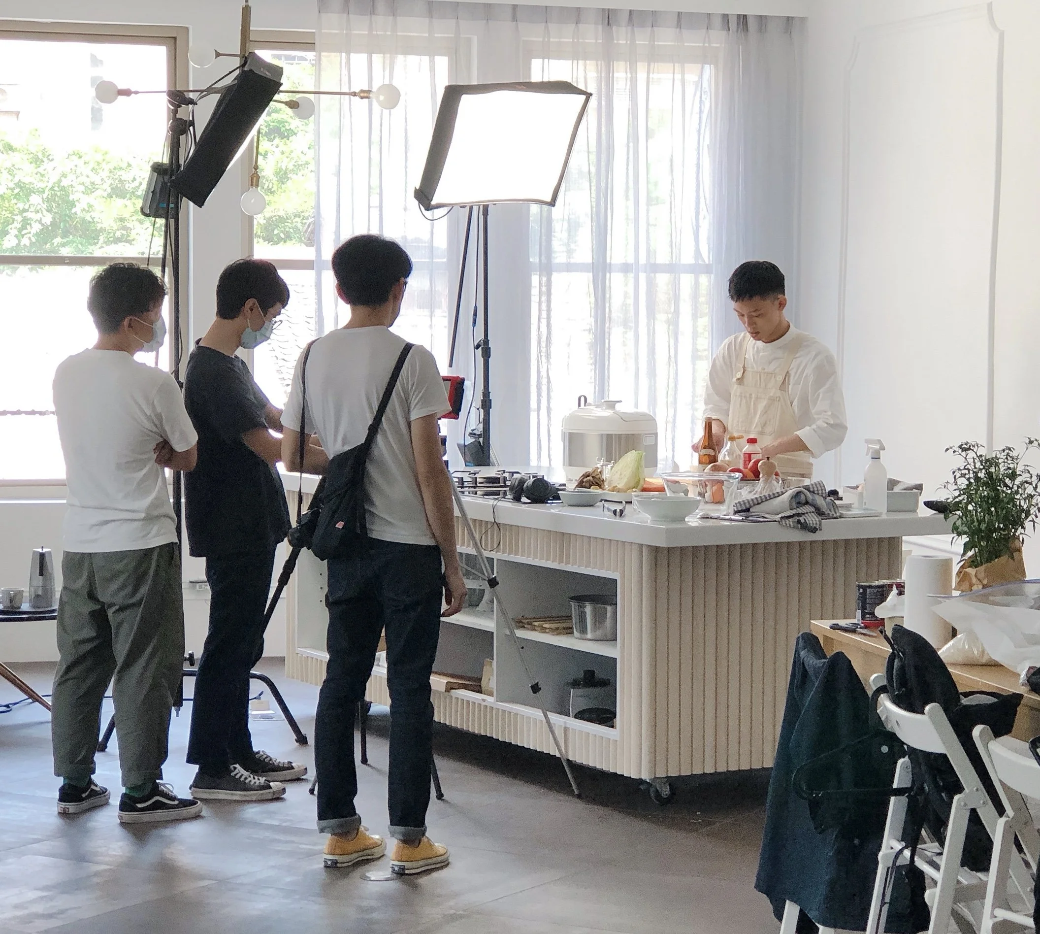 A daytime indoor photo of a cooking show set with three people filming a chef who is preparing food at a countertop. The chef stands behind a kitchen island with various ingredients and cooking utensils. Lighting equipment and a camera are set up to 