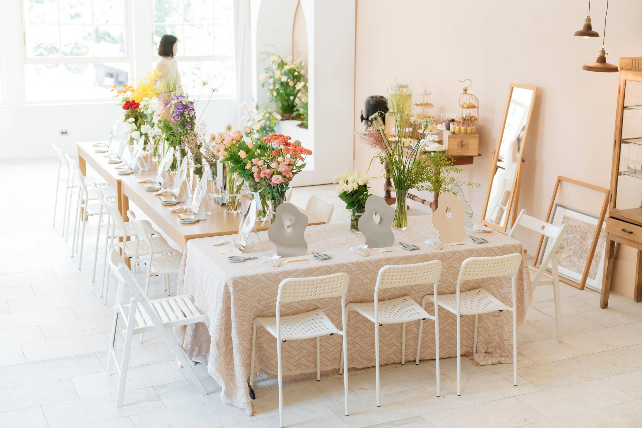 A decorated dining room with a long table and chairs, filled with colorful flowers in vases, set for a meal, with large windows letting in natural light.