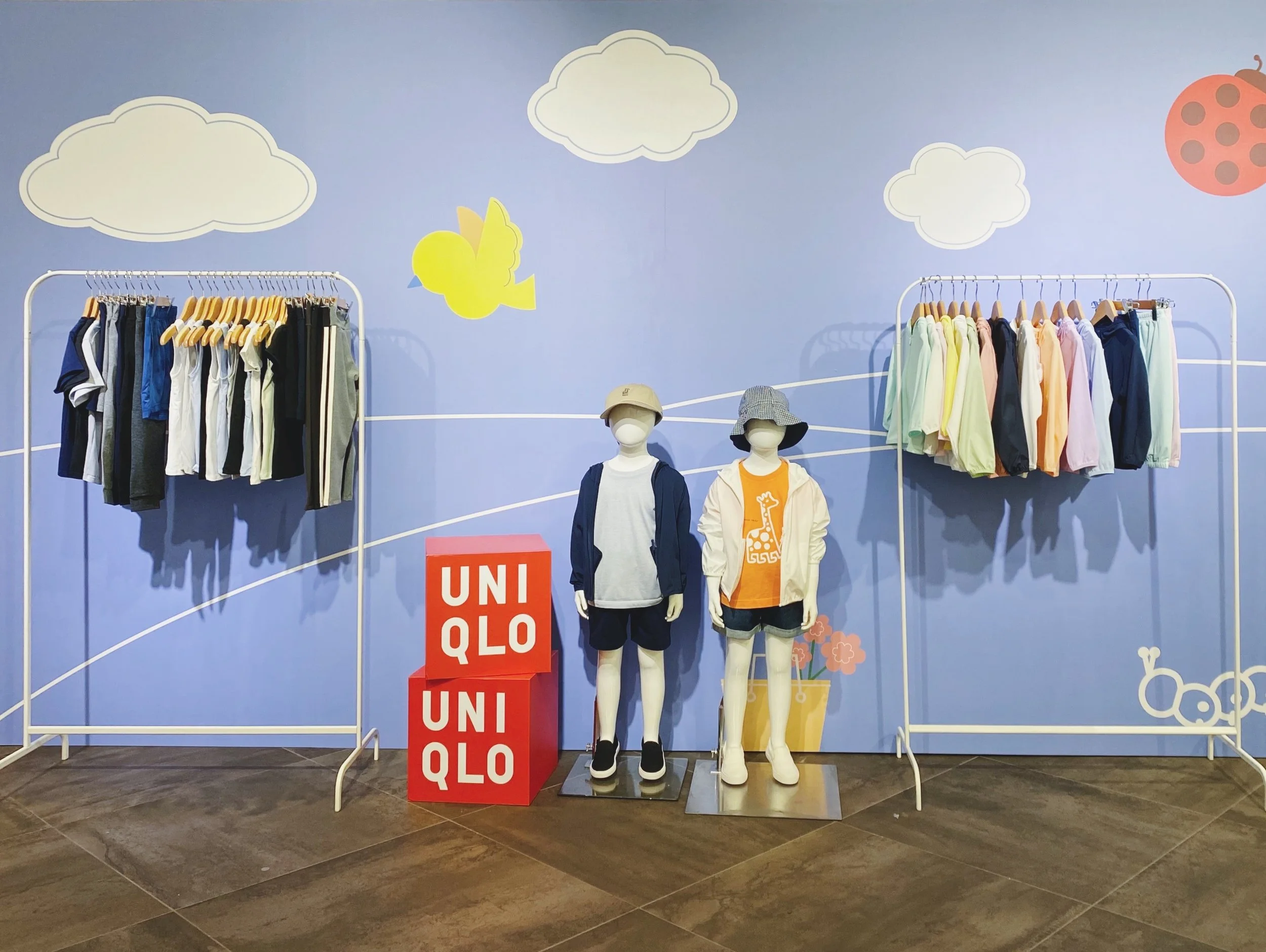 Kids' clothing display in a store with a blue wall decorated with clouds, a bird, and a ladybug. Two mannequins wear casual outfits, and two racks hold colorful children's clothes. Red blocks with white UNIQLO logo are on the floor.