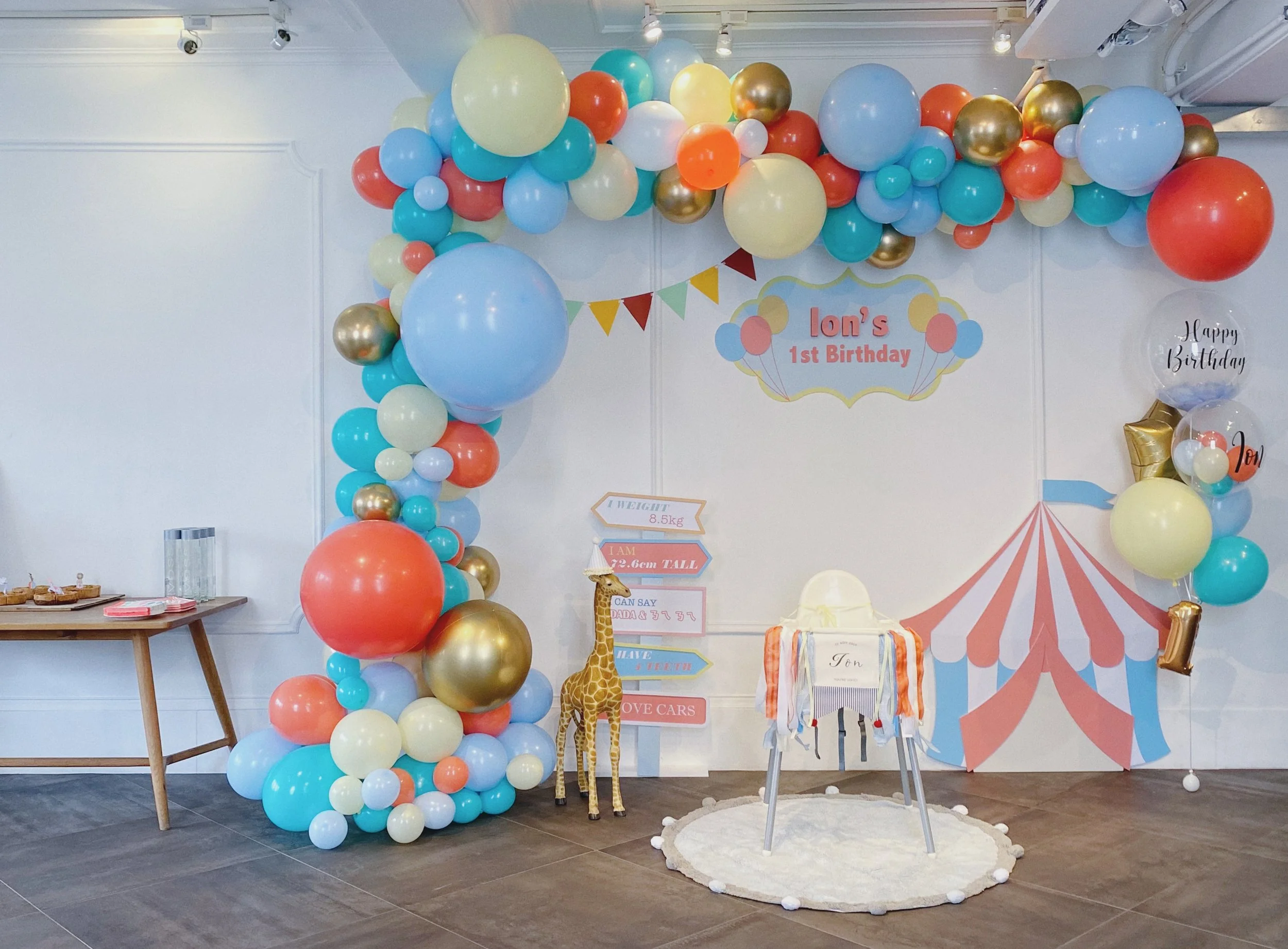 Colorful balloons decorating a birthday party setup with a sign that reads "Ion's 1st Birthday" and a small giraffe toy on the floor. There is a table with cupcakes and a backdrop with a circus tent illustration.