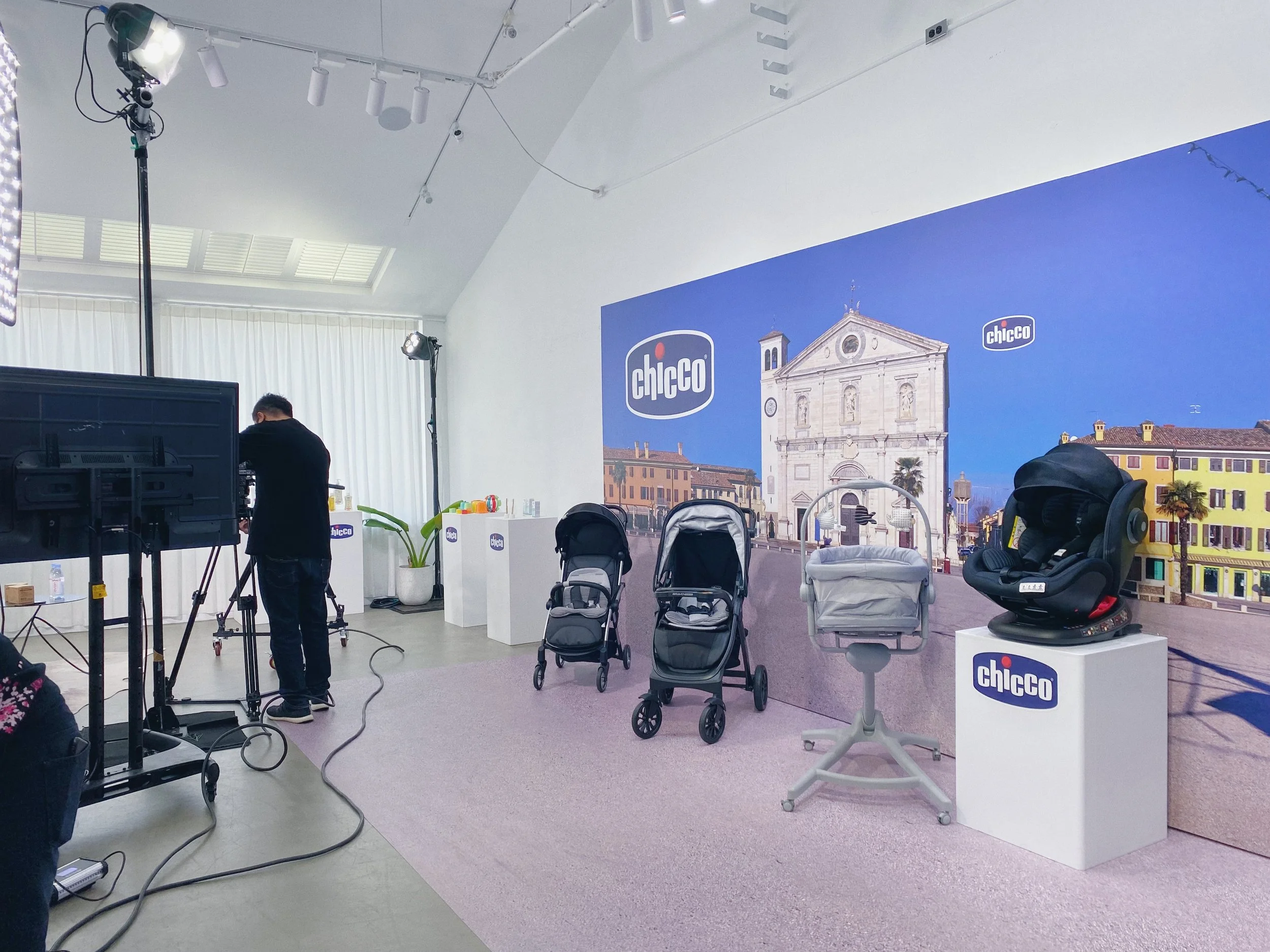 Display setup with baby strollers, car seats, and a backdrop featuring a cityscape with a church and colorful buildings, branded with Chickco logos, in a well-lit room with studio lighting.