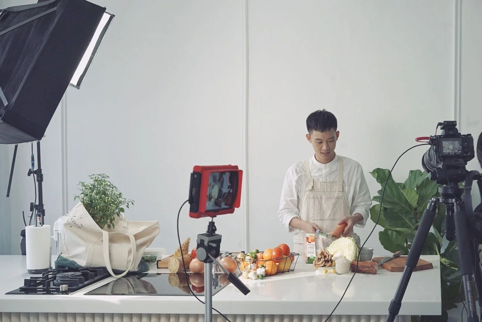Person in a white shirt and apron preparing vegetables in a filming studio with cooking equipment, plants, and produce on the countertop.