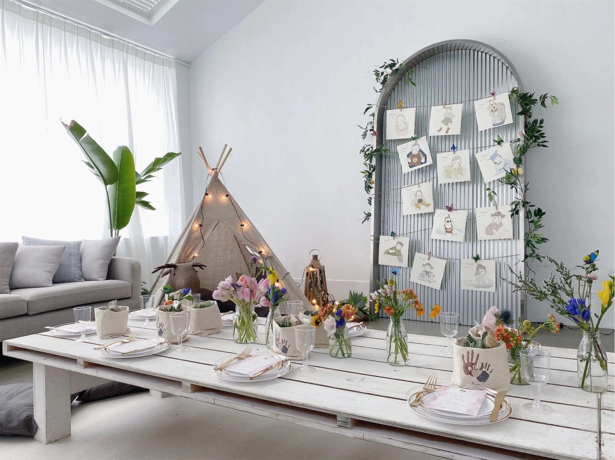 A cozy indoor space decorated with a white wooden dining table, colorful flowers in glass vases, a beige teepee with fairy lights, a neutral-colored sofa, and a large arched metal backdrop decorated with children's drawings clipped to strings and sur