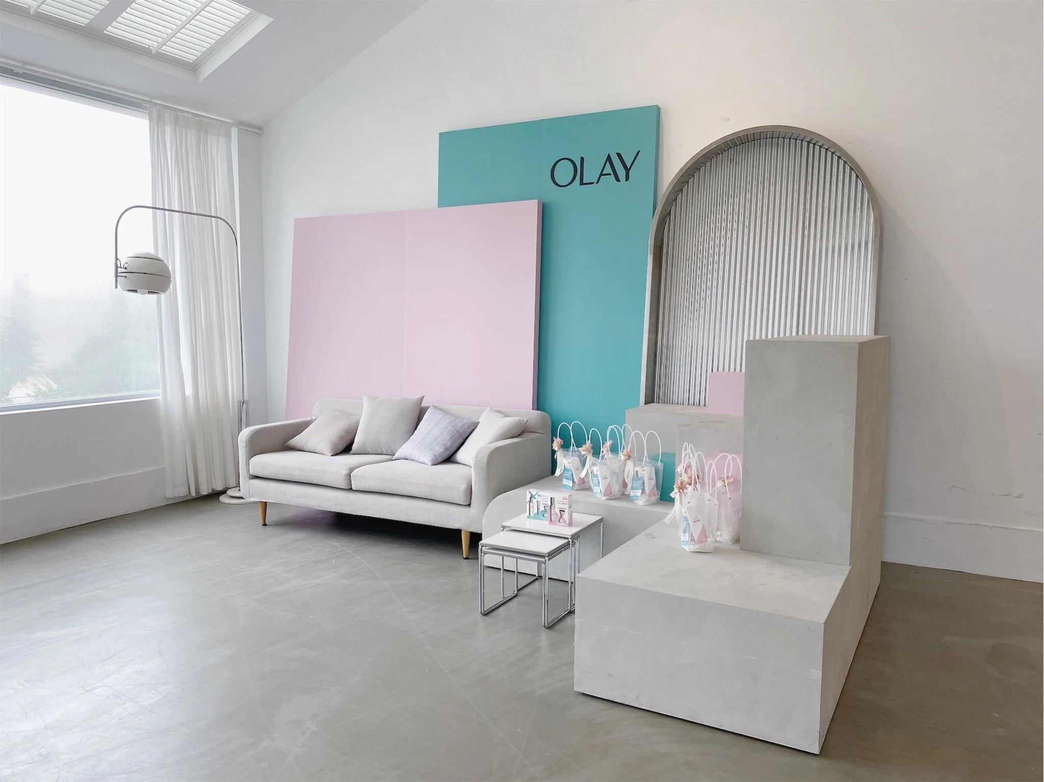 Interior of a bright room with a white sofa, pastel-colored backdrops, gift bags on a step, and a sign that says 'OLAY'.