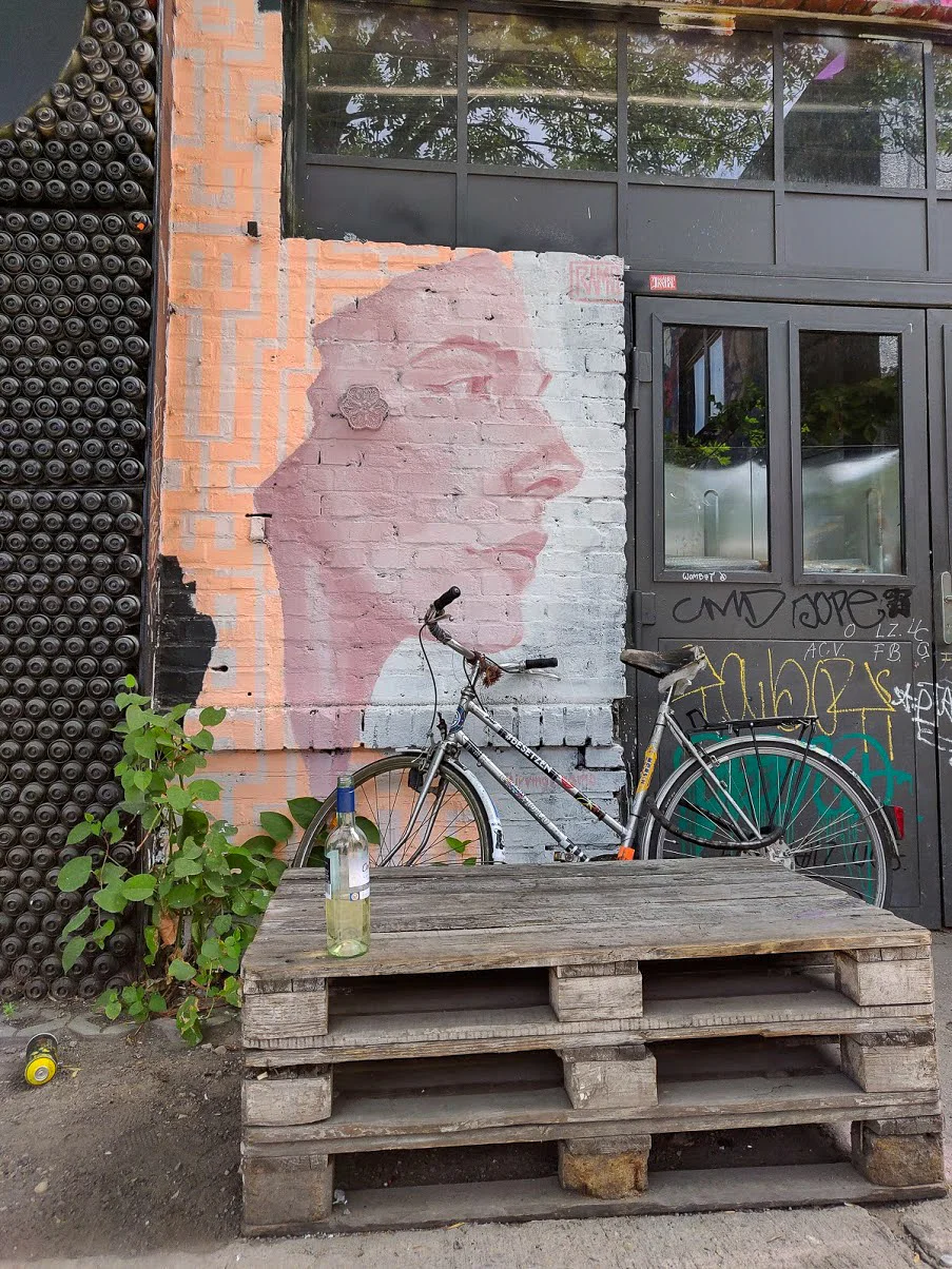 Den här bilden är en sammanfattning av Berlin: gatukonst, graffiti, en vägg gjord av gamla sprayflaskor, en sprayflaska, en lastpall, en cykel och en bortglömd vinflaska.