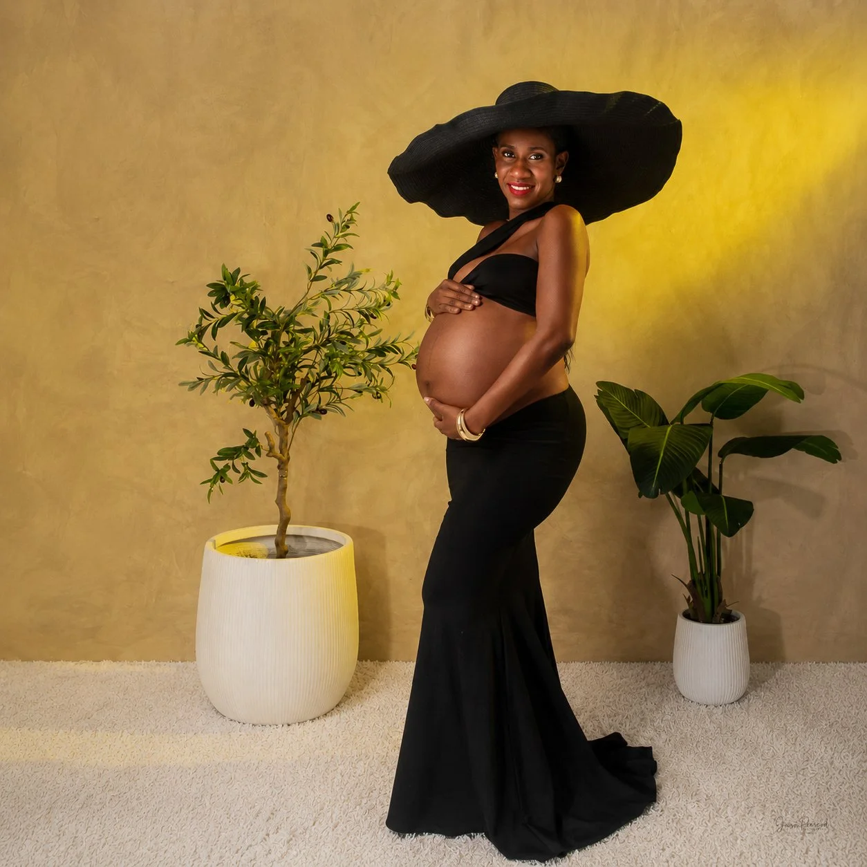 Pregnant woman wearing a large black hat and black outfit, standing next to potted plants against a yellow background.