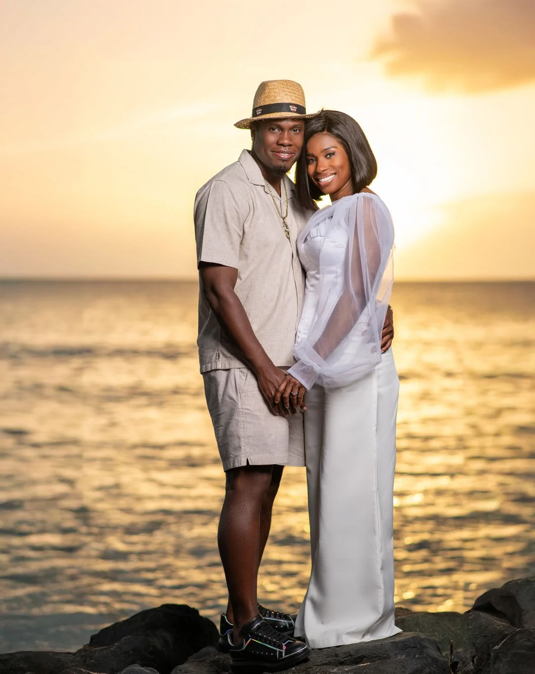A couple standing on rocks at the beach during sunset, holding hands and smiling at the camera. The man is wearing a straw hat, light beige shirt, and shorts. The woman is in a white dress with sheer sleeves.