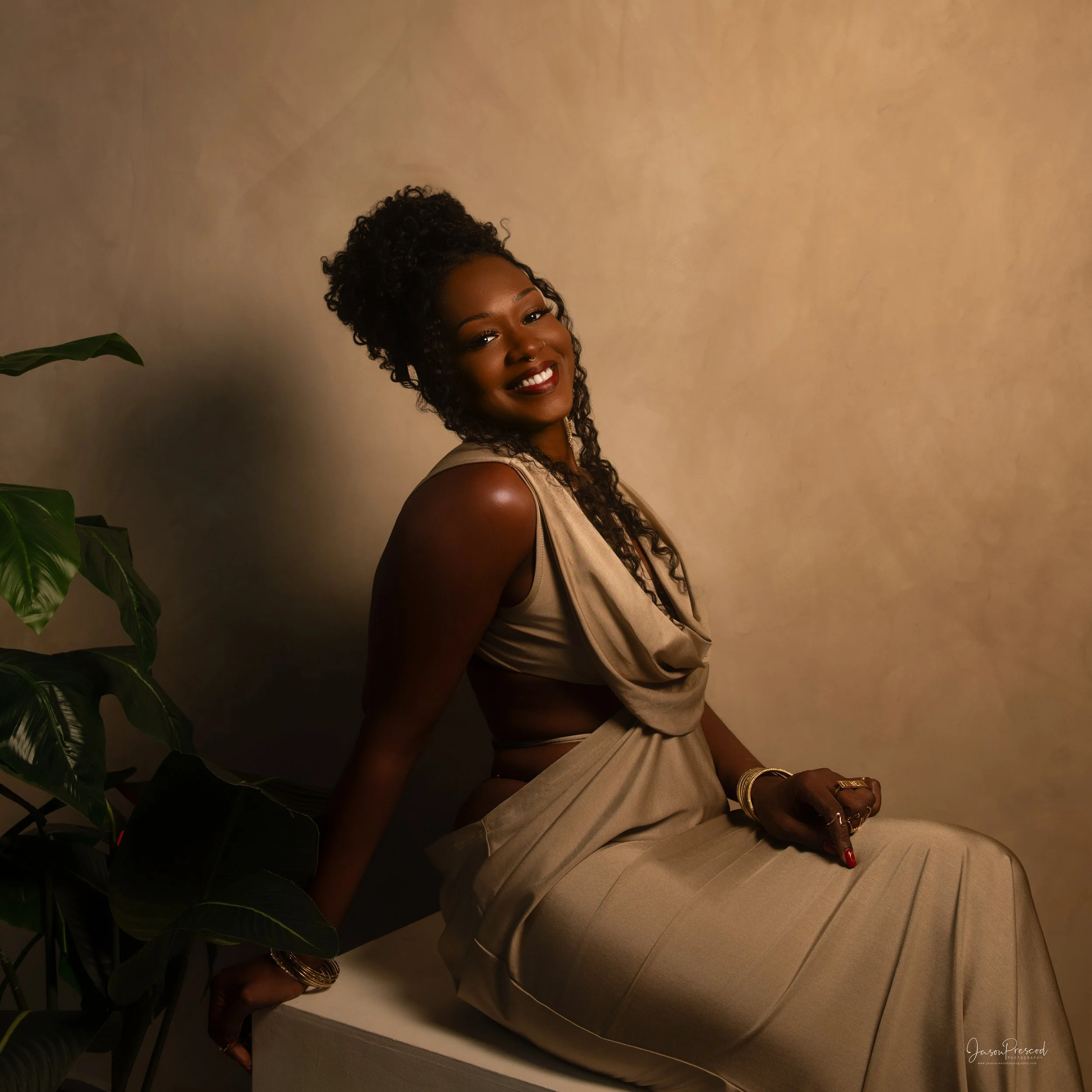 A woman with curly black hair tied up, smiling, wearing gold jewelry and a beige outfit, posing against a plain beige wall with a green plant partially visible on the left side of the image.