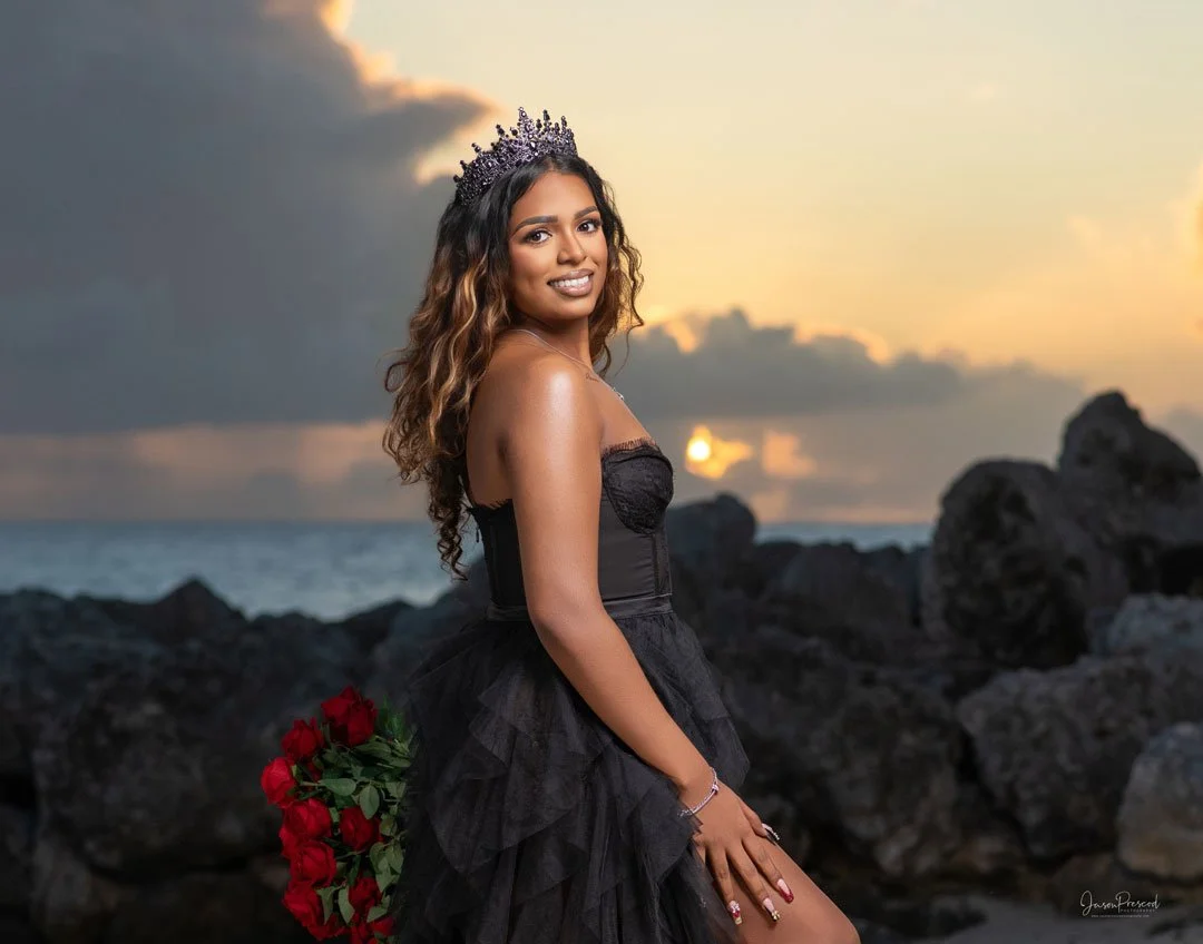 A young woman in a black dress and tiara holding red roses on a rocky beach at sunset.