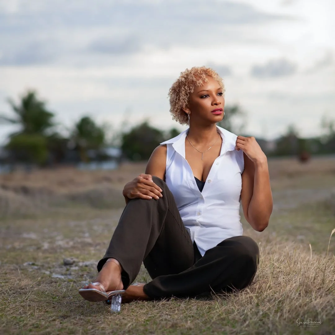 A woman with curly blonde hair sitting on the grass outdoors, wearing a sleeveless white blouse and black pants, looking thoughtfully into the distance.