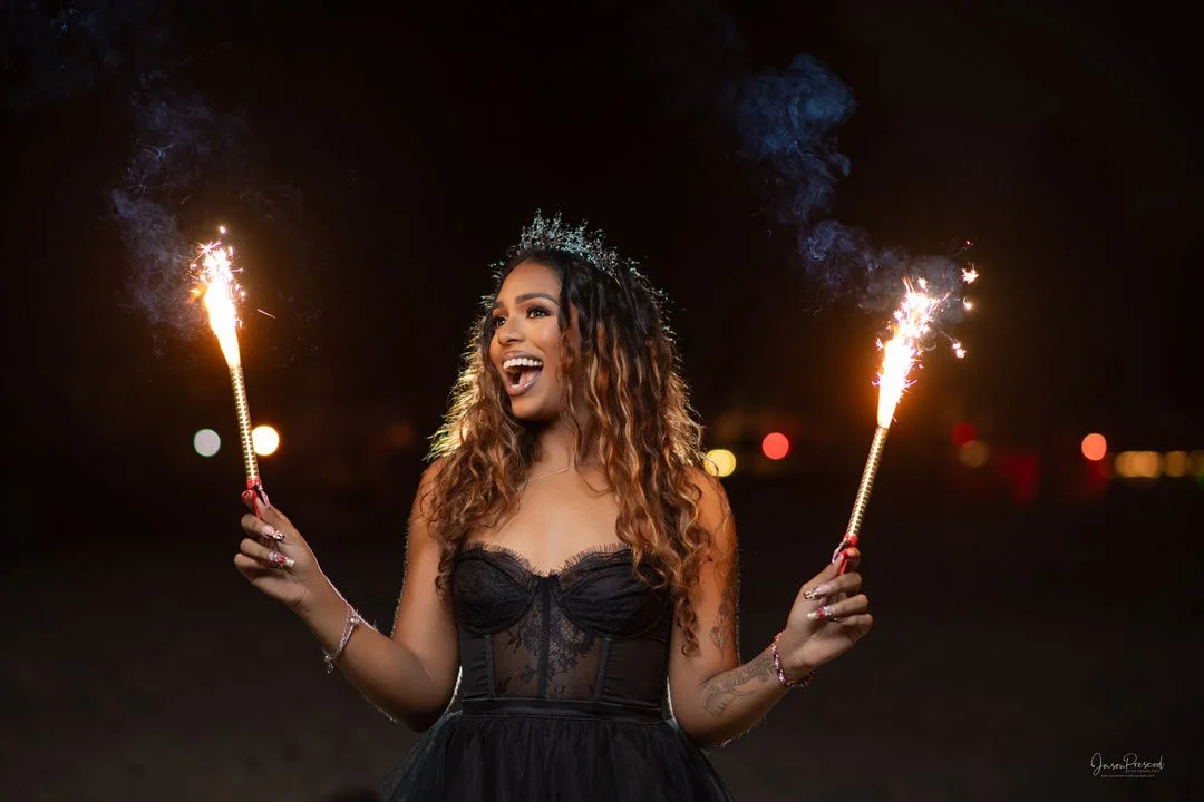 Woman with curly hair wearing a black dress and tiara, holding sparklers in both hands, smiling and celebrating at night.