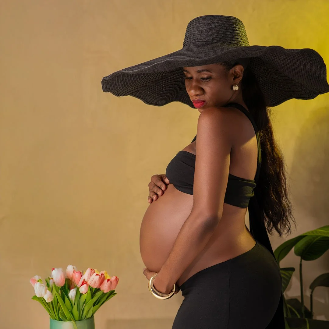 A pregnant woman wearing a large black sun hat, black top, and black skirt, standing indoors near a yellow wall and a green potted plant, holding her baby bump, with a bouquet of pink and white tulips on a table nearby.