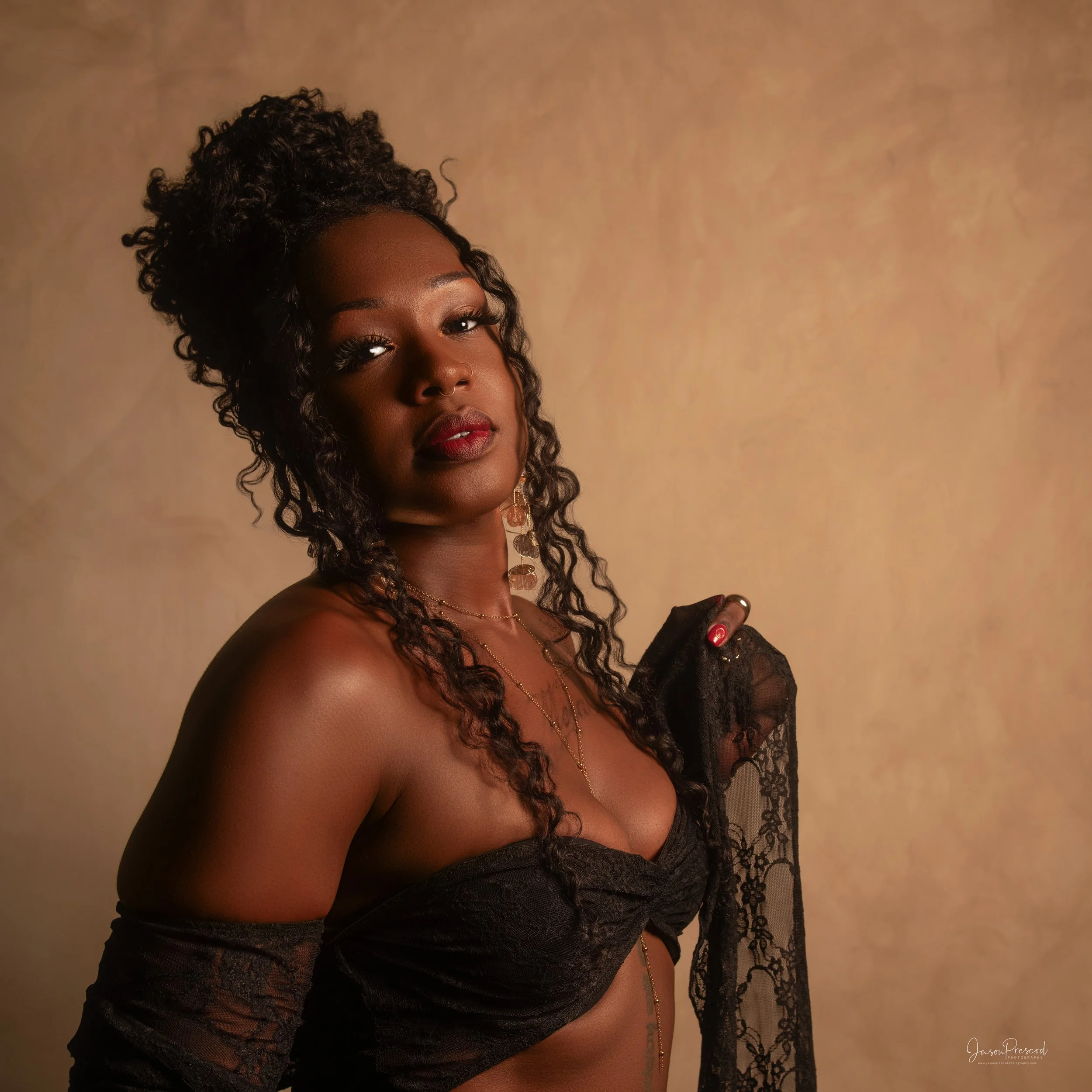 A close-up portrait of a woman with curly hair wearing a black lace off-shoulder top and layered gold jewelry against a beige background.